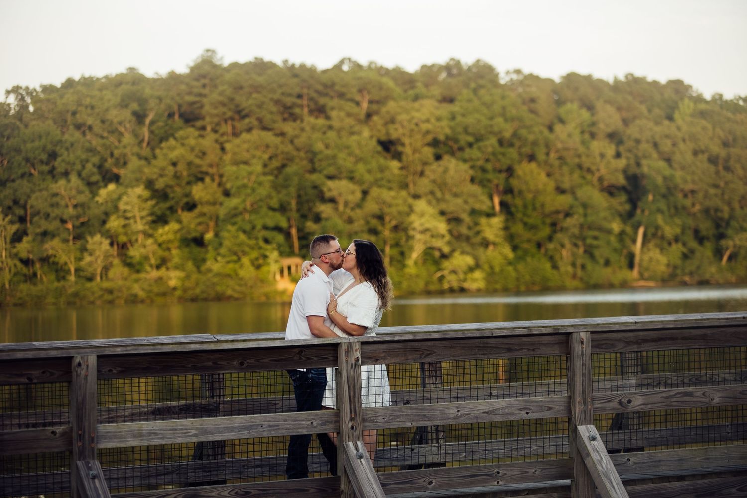 A fishing pier with metal railings extends over a serene lake surrounded by dense green forest in the afternoon light.