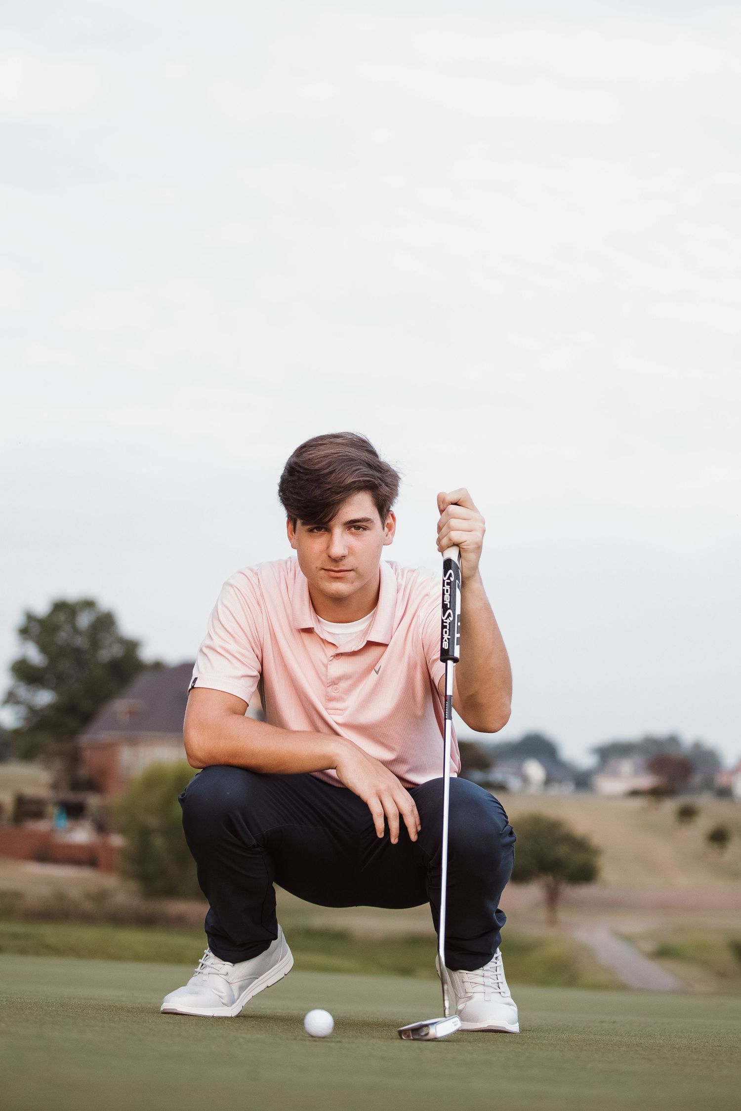 Golfer in pink polo shirt crouches on green to read putting line on golf course.