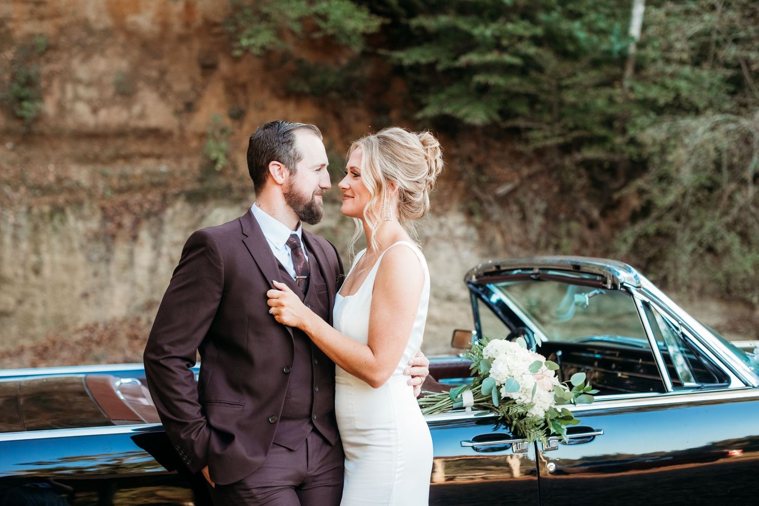 Newlywed couple shares romantic moments in front of vintage blue car during outdoor forest wedding photoshoot.