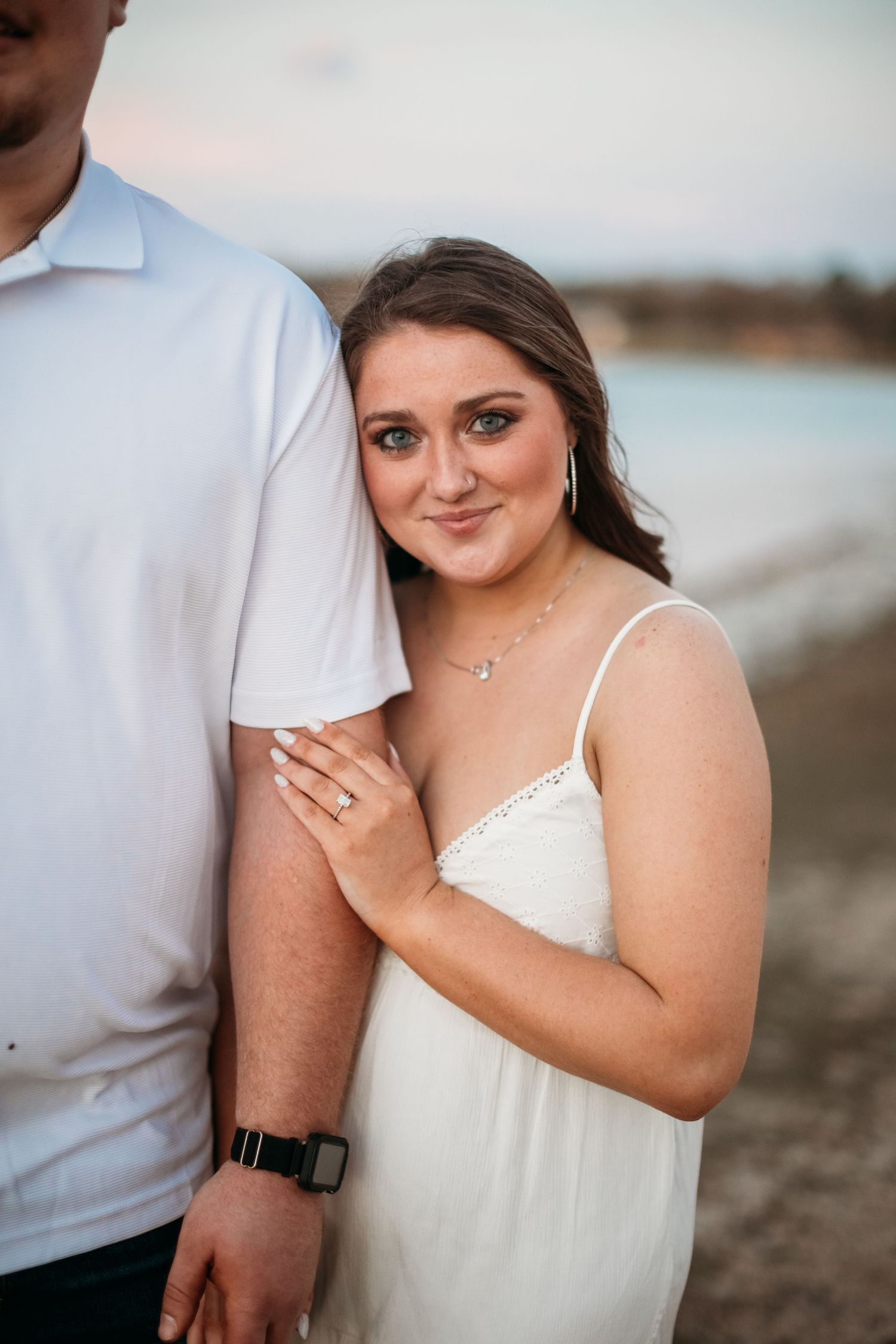 Woman in white dress leans against man in white polo on beach at sunset, smiling at camera.