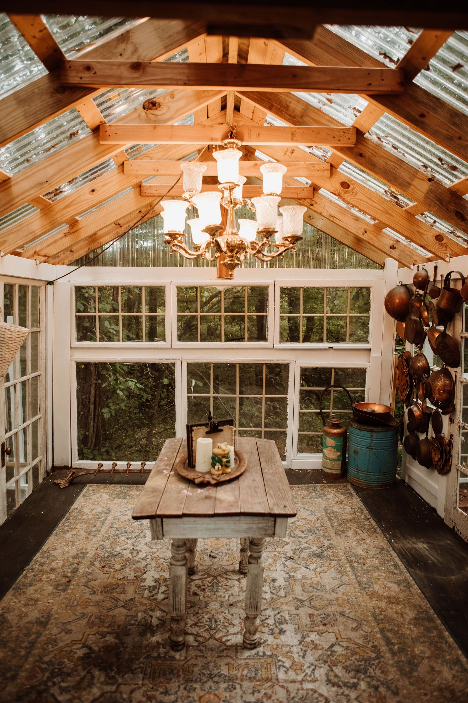 Rustic greenhouse room with a glass roof, wooden beams, vintage chandelier, farmhouse table, patterned rug and hanging copper pots.