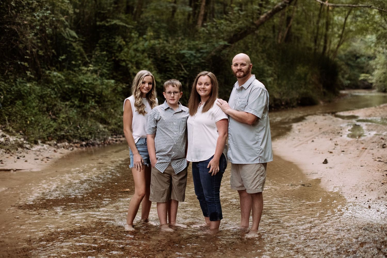 A family of four poses together on a sandy creek bed surrounded by lush green forest.