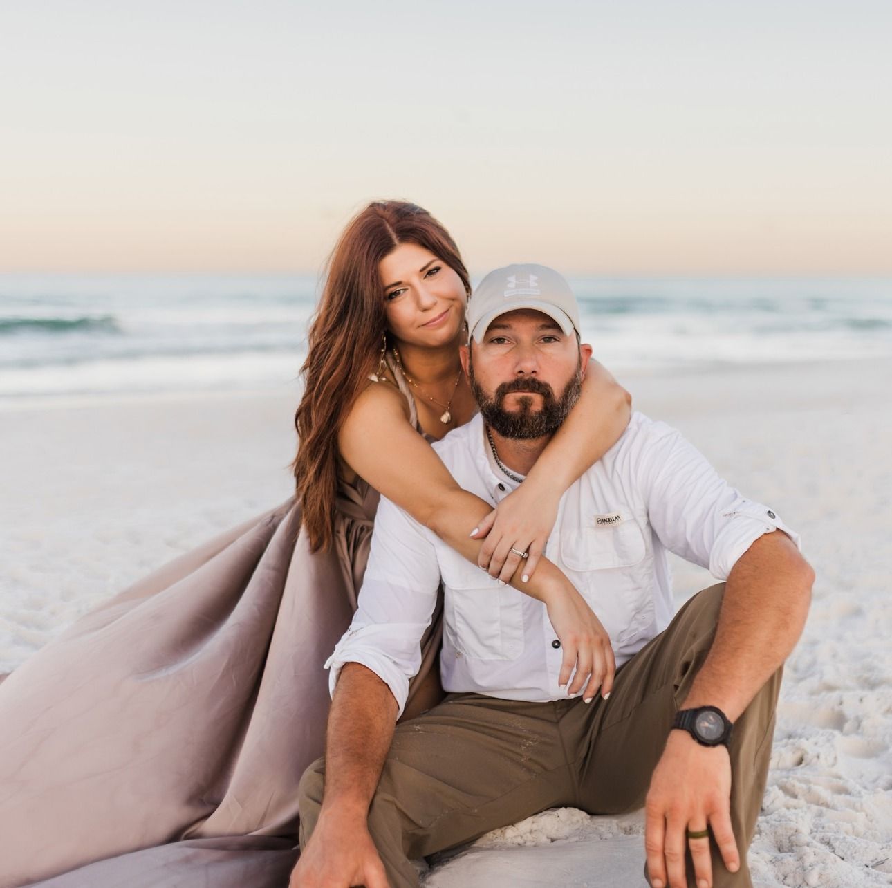 Couple embraces on sandy beach at sunset with soft pink sky and ocean waves in background.