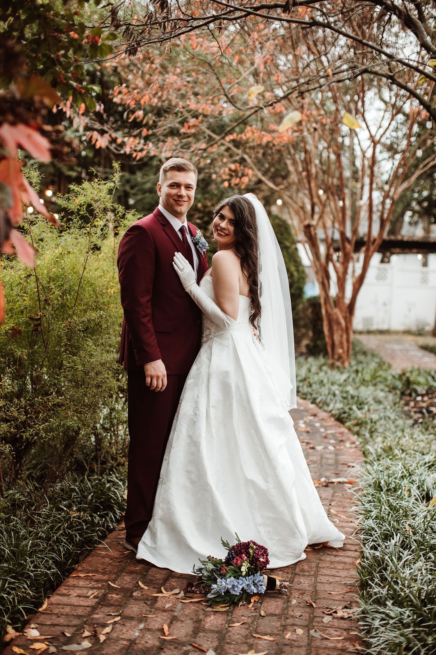Bride in white gown and groom in burgundy suit pose together on brick path surrounded by autumn foliage.