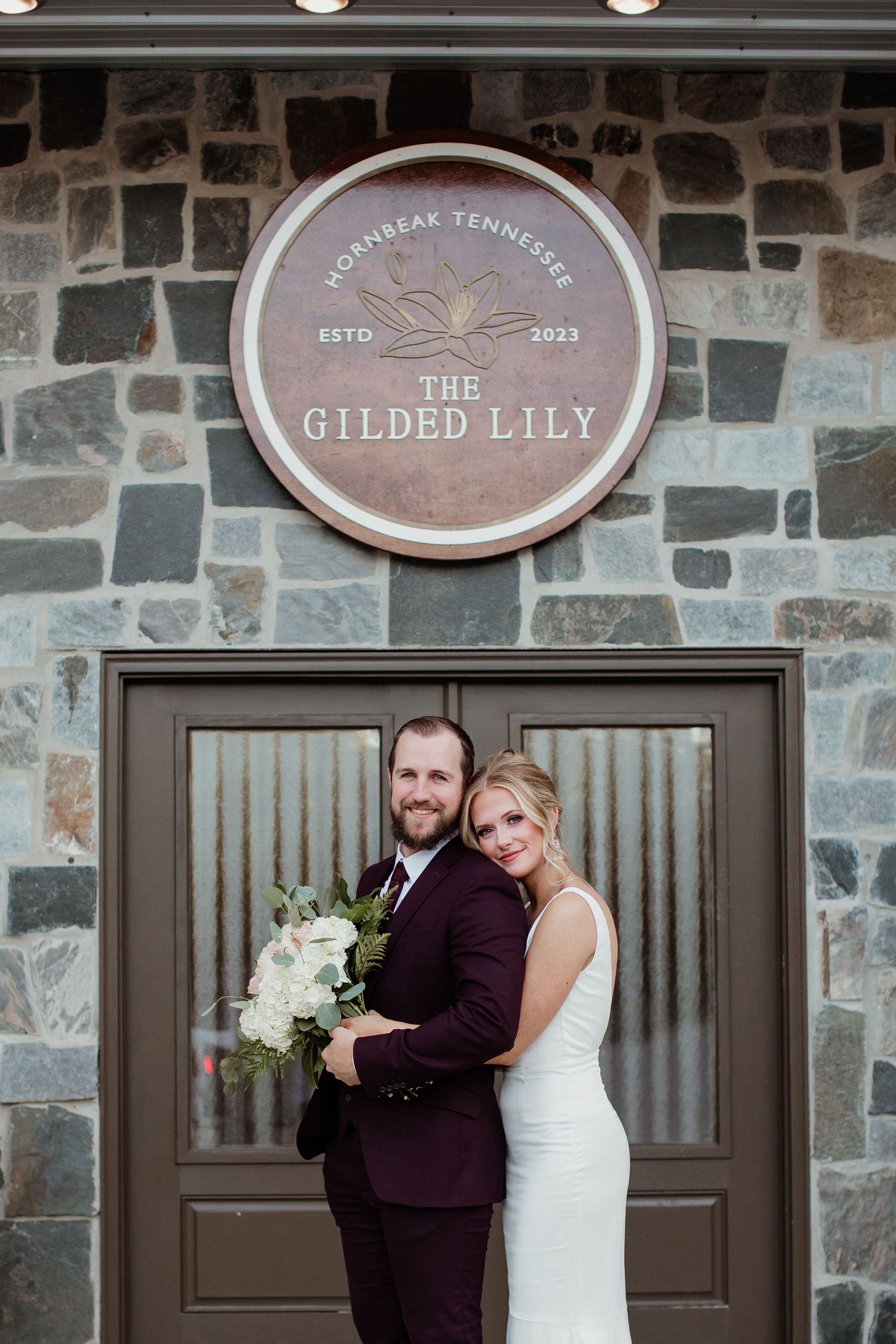 A romantic wedding couple poses outside The Gilded Lily venue with stone walls and elegant brown doors.