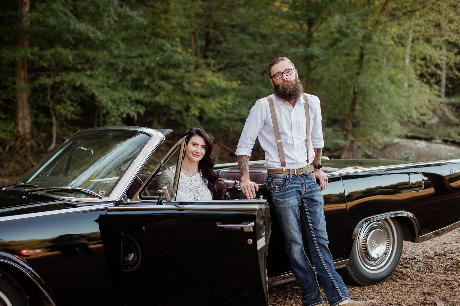 A couple stands near a classic black car in a wooded setting during an autumn photo session.