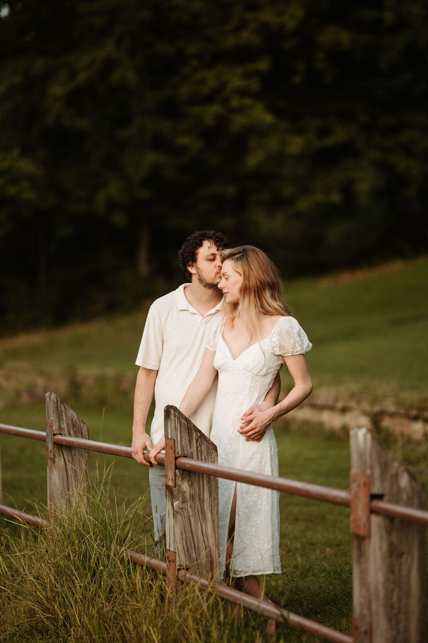 A romantic couple shares a tender moment while leaning against a rustic wooden fence in a grassy field at sunset.