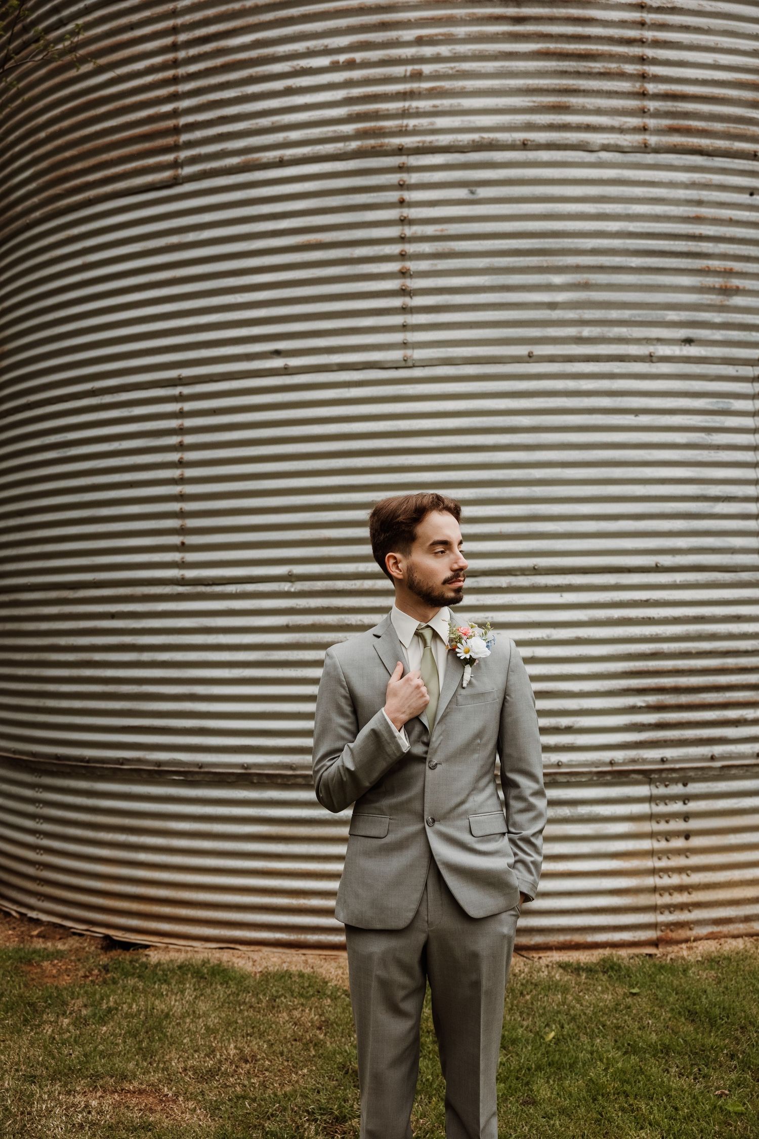 Groom in gray suit with boutonniere stands before rustic corrugated metal grain silo at outdoor wedding.