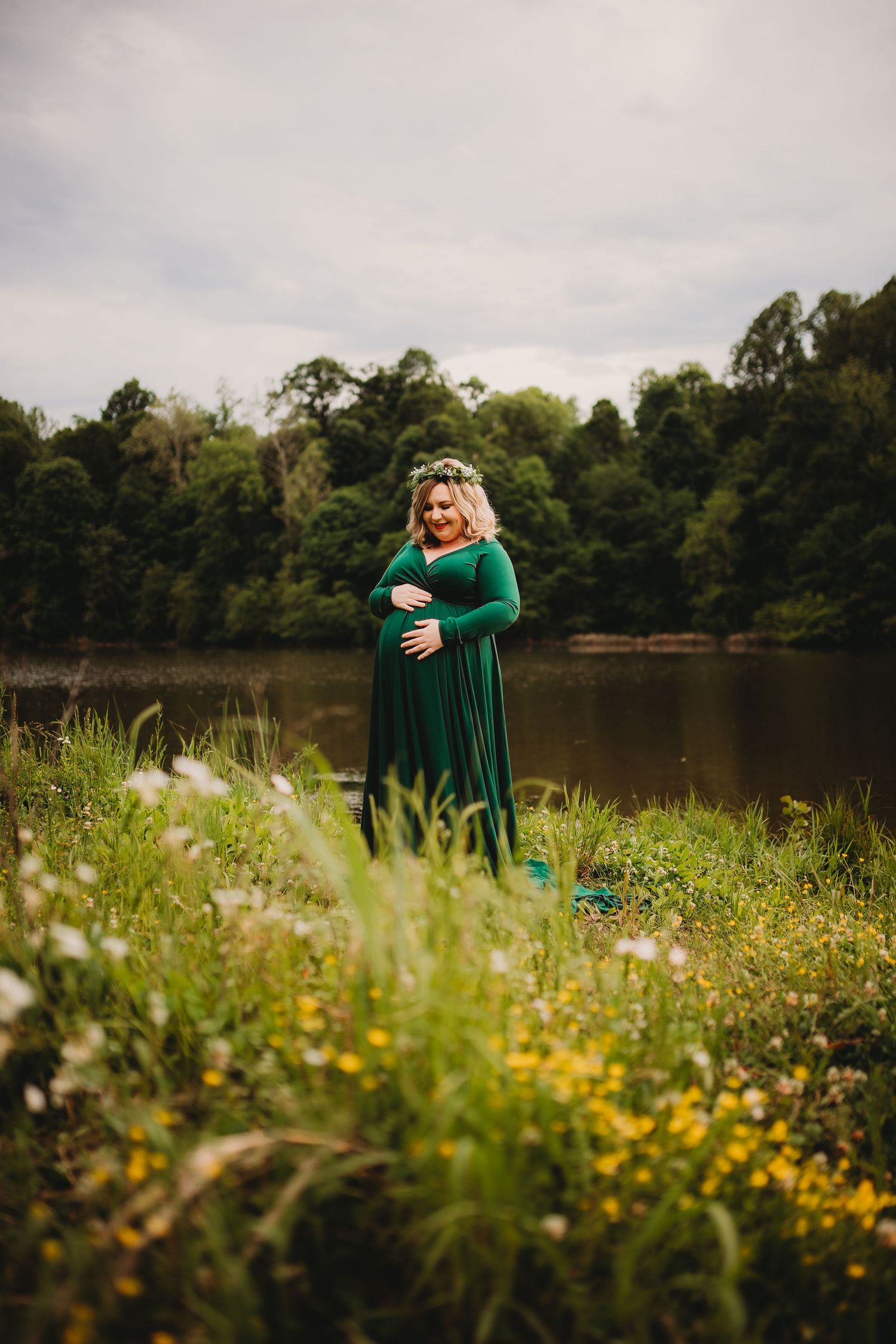 A person in an emerald green dress poses near a tranquil lake surrounded by wildflowers.