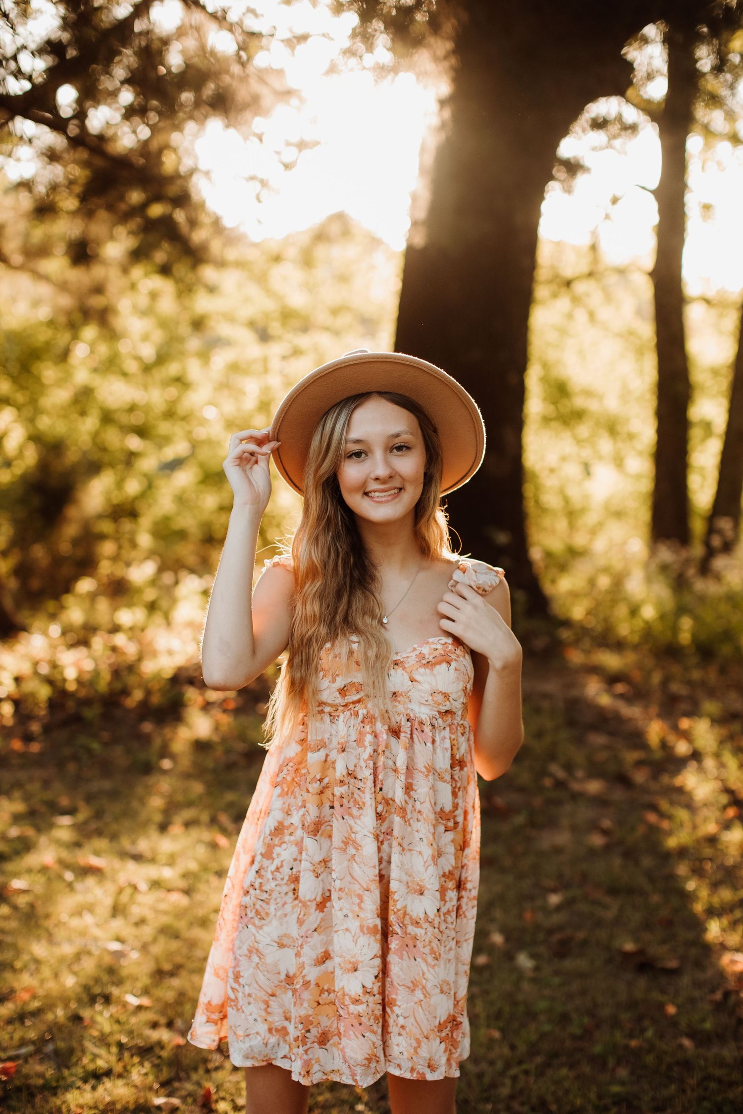 Floral sundress and wide-brimmed hat styled in golden sunset lighting through trees in forest setting.