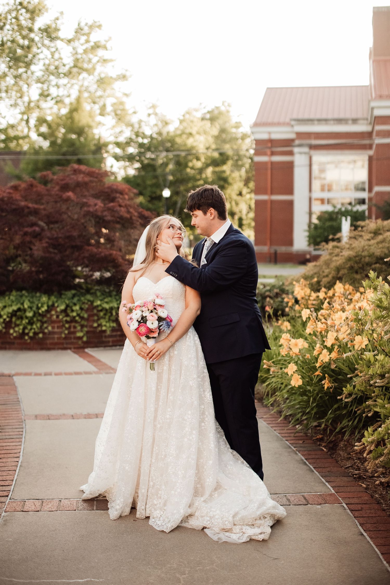 A bride and groom share an intimate moment on a brick pathway surrounded by lush gardens and brick buildings.