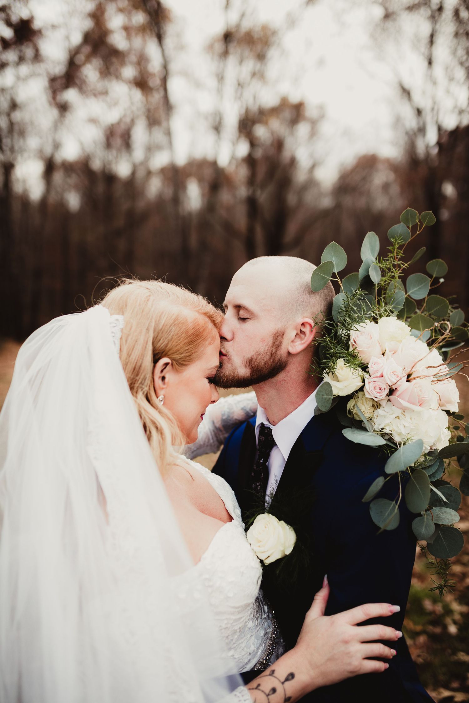 Romantic wedding couple shares a tender kiss outdoors in autumn with white floral bouquet and greenery.
