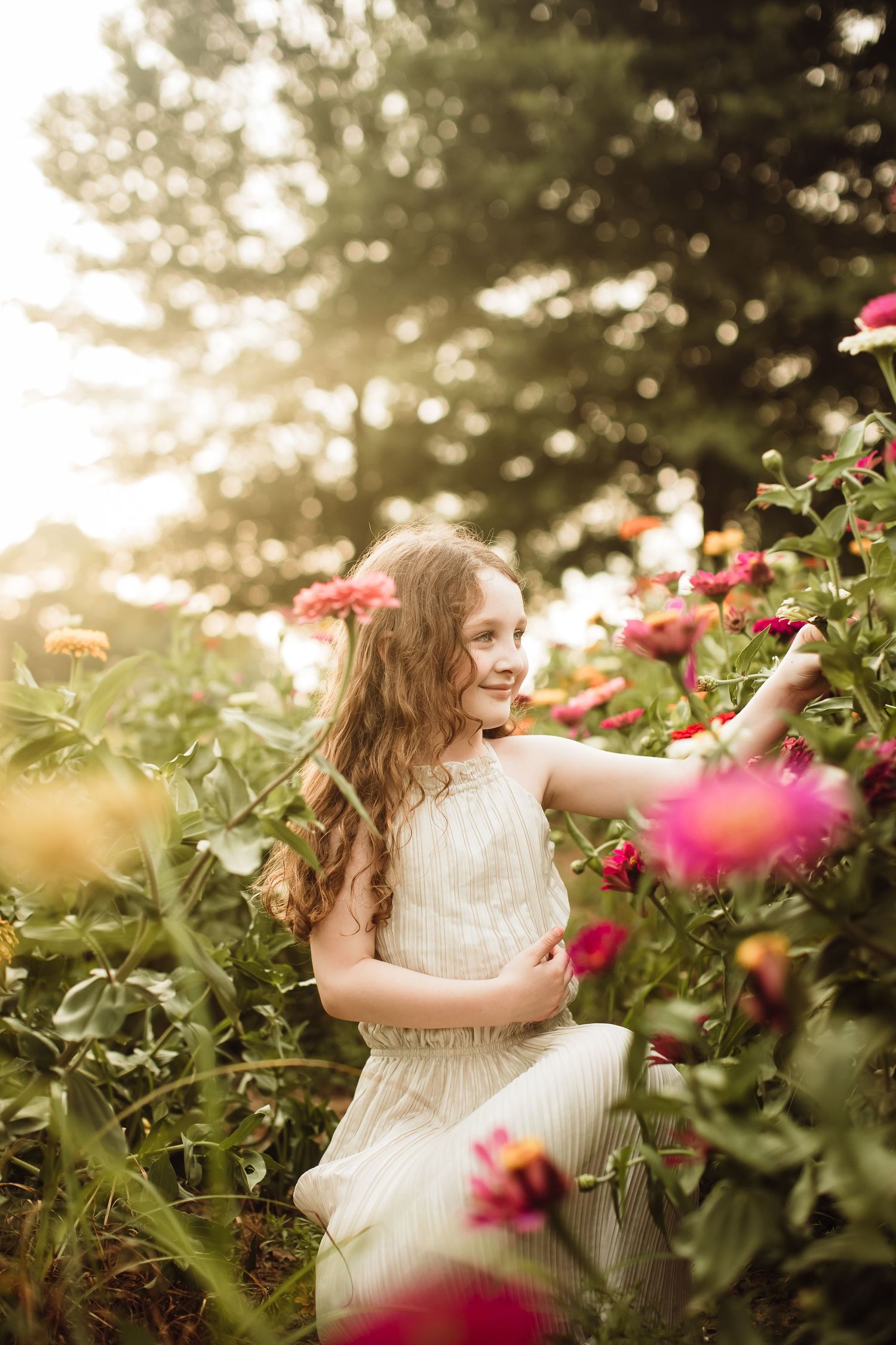 Young girl in white dress reaching for pink flowers in a sunlit garden, golden hour light filtering through trees.