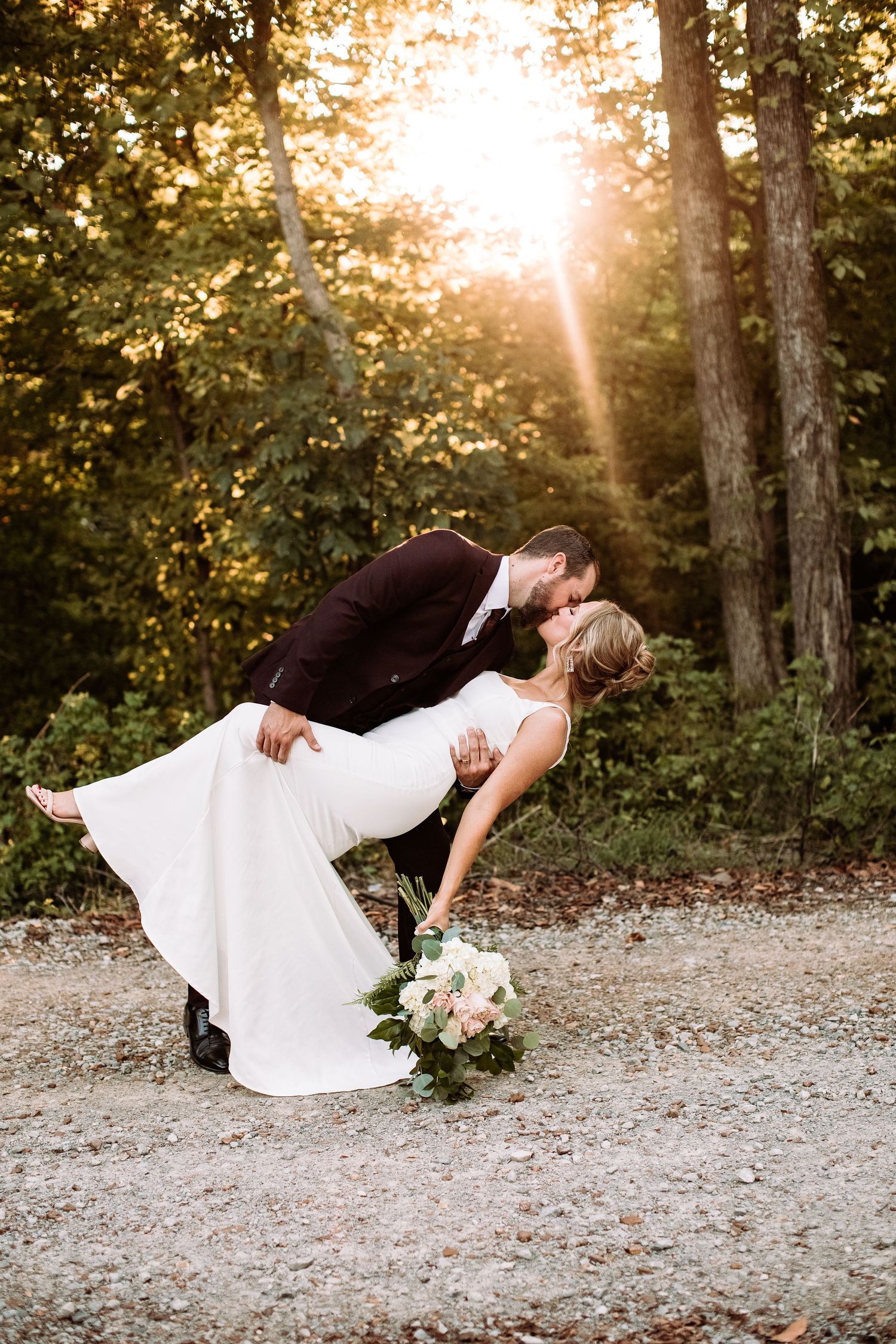 A couple in wedding attire performs a romantic dip pose in a sunlit forest setting at sunset.