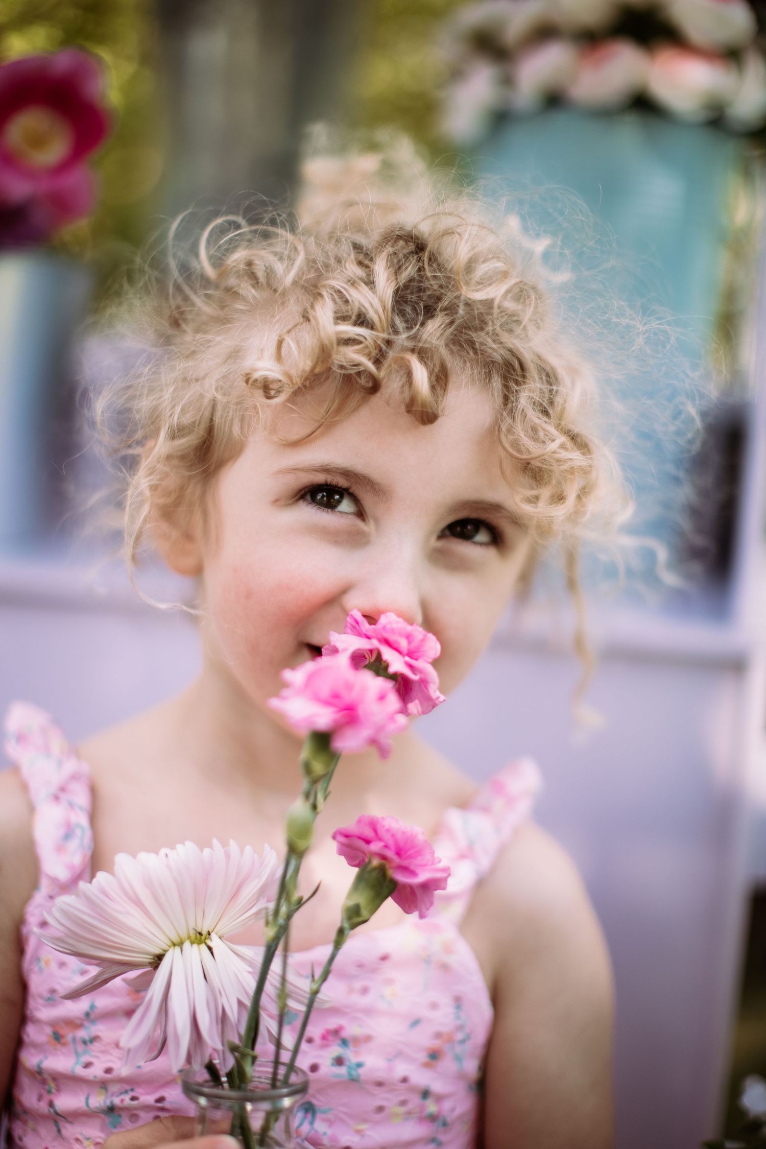 Child with curly blonde hair smelling pink flowers in springtime garden setting.