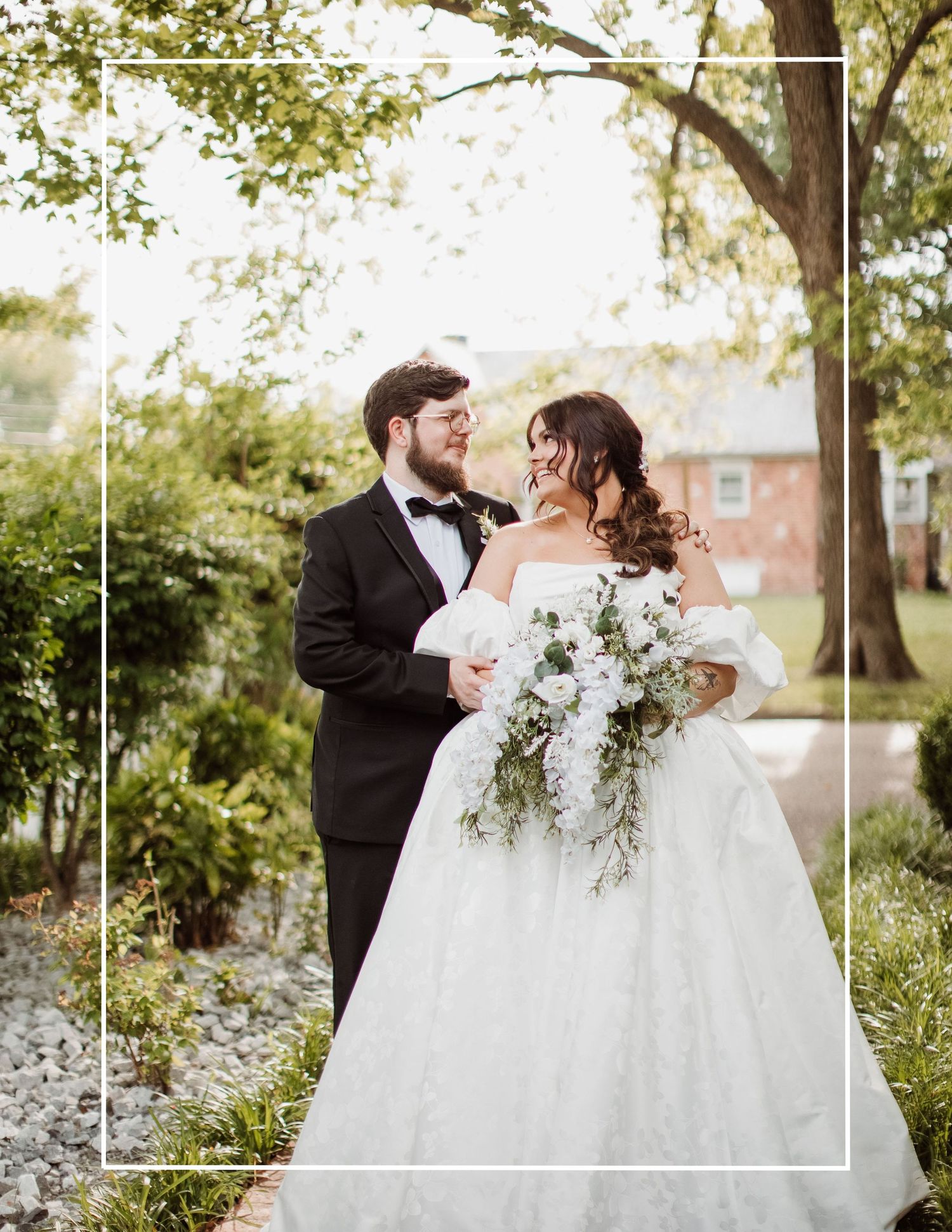 A romantic wedding couple shares a tender moment outdoors surrounded by lush greenery and white floral arrangements.