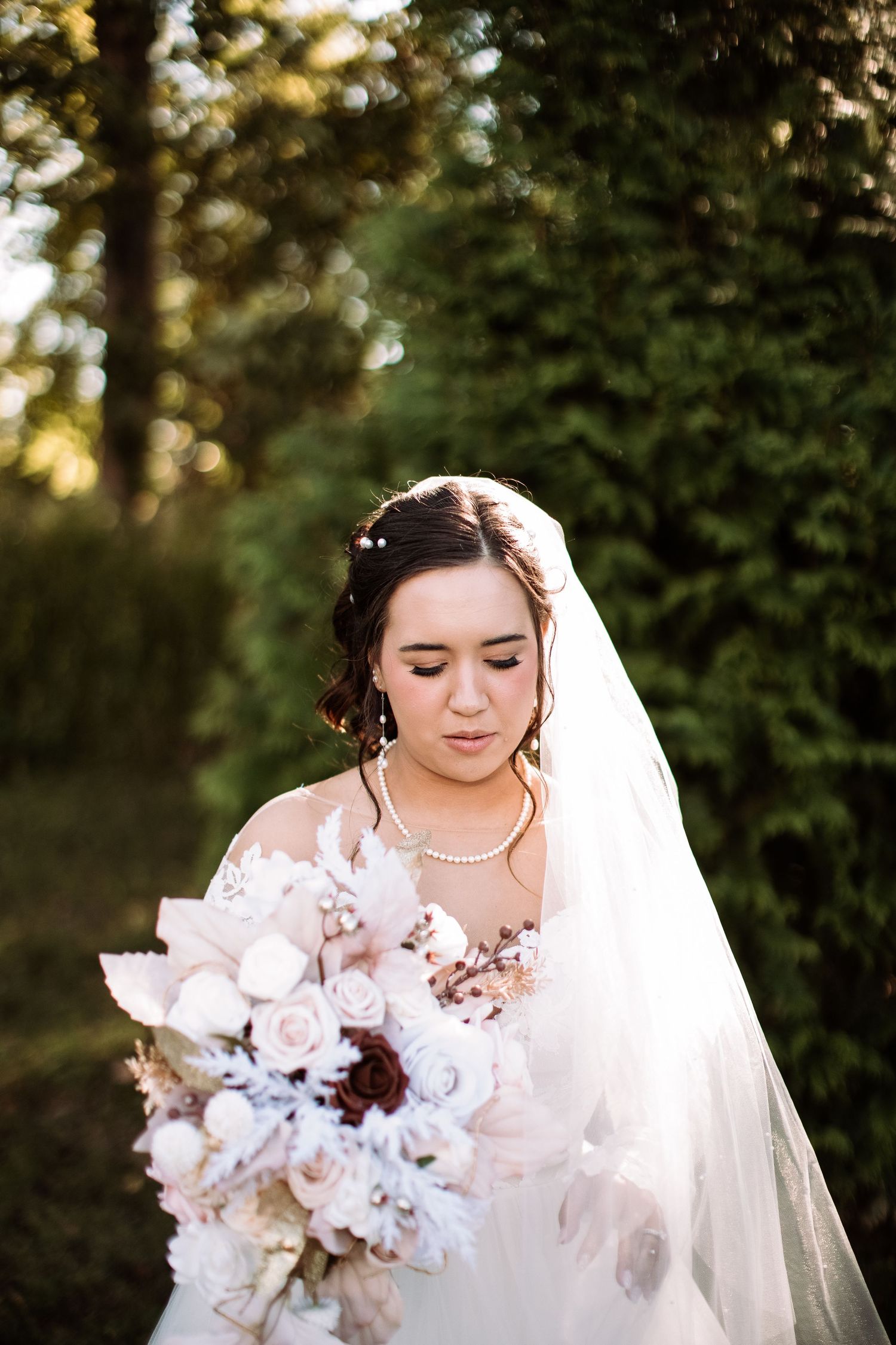 A bride in a white wedding dress holds a soft pink and white floral bouquet against a blurred outdoor garden background.