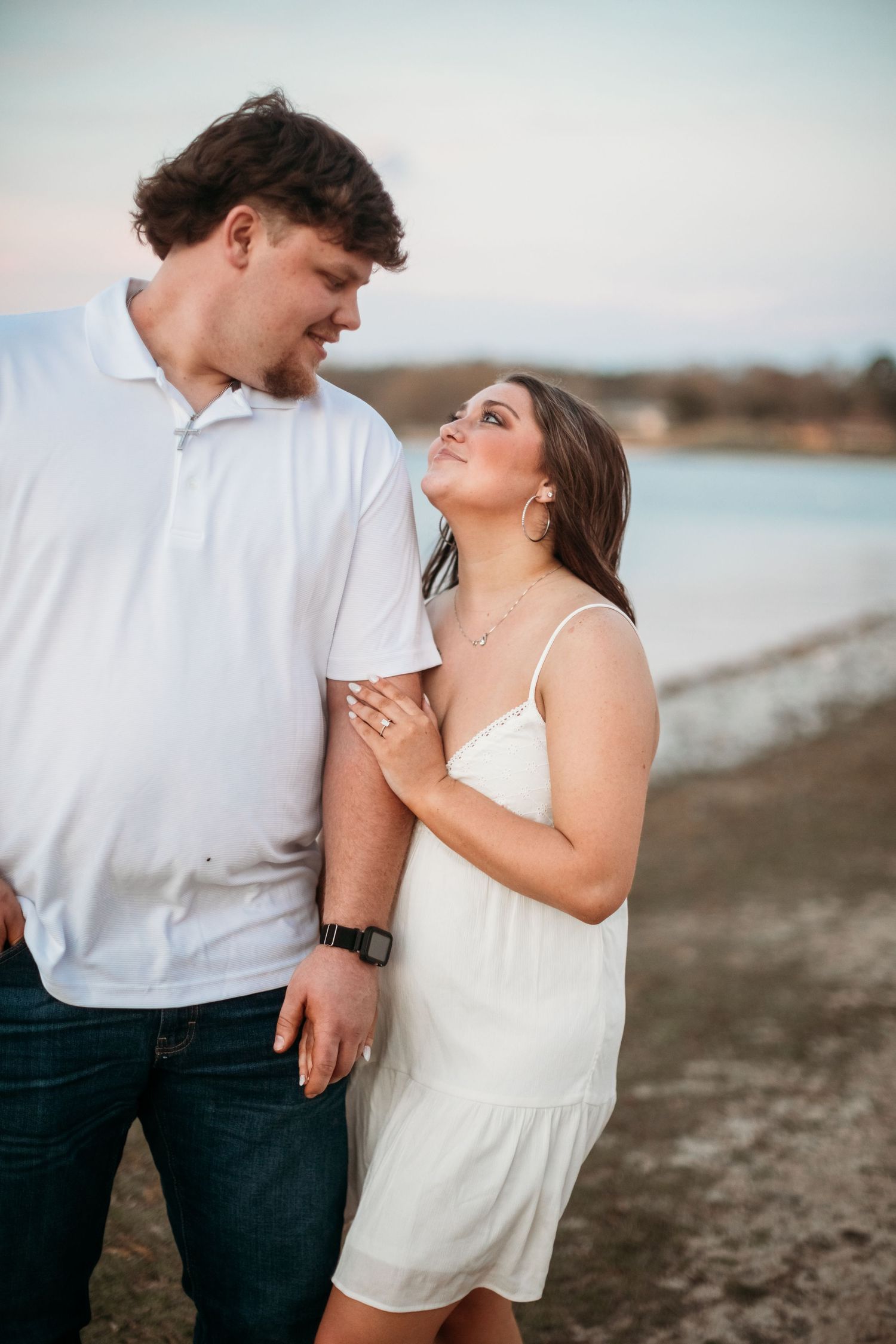 Couple in white outfits sharing a romantic moment near a lake at sunset, woman in white dress gazing up at man.