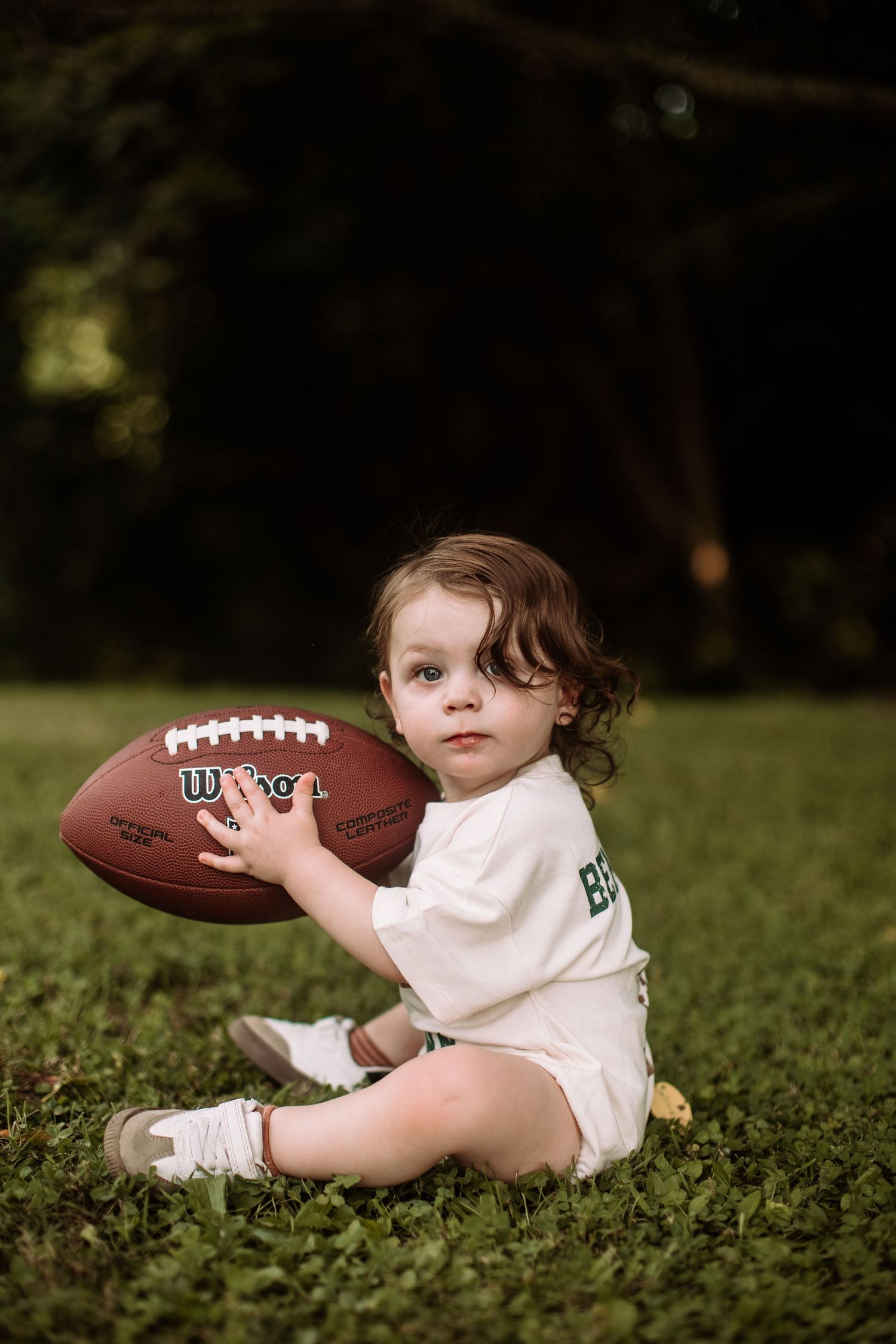 Small child in white outfit sits on grass holding Wilson football outdoors.