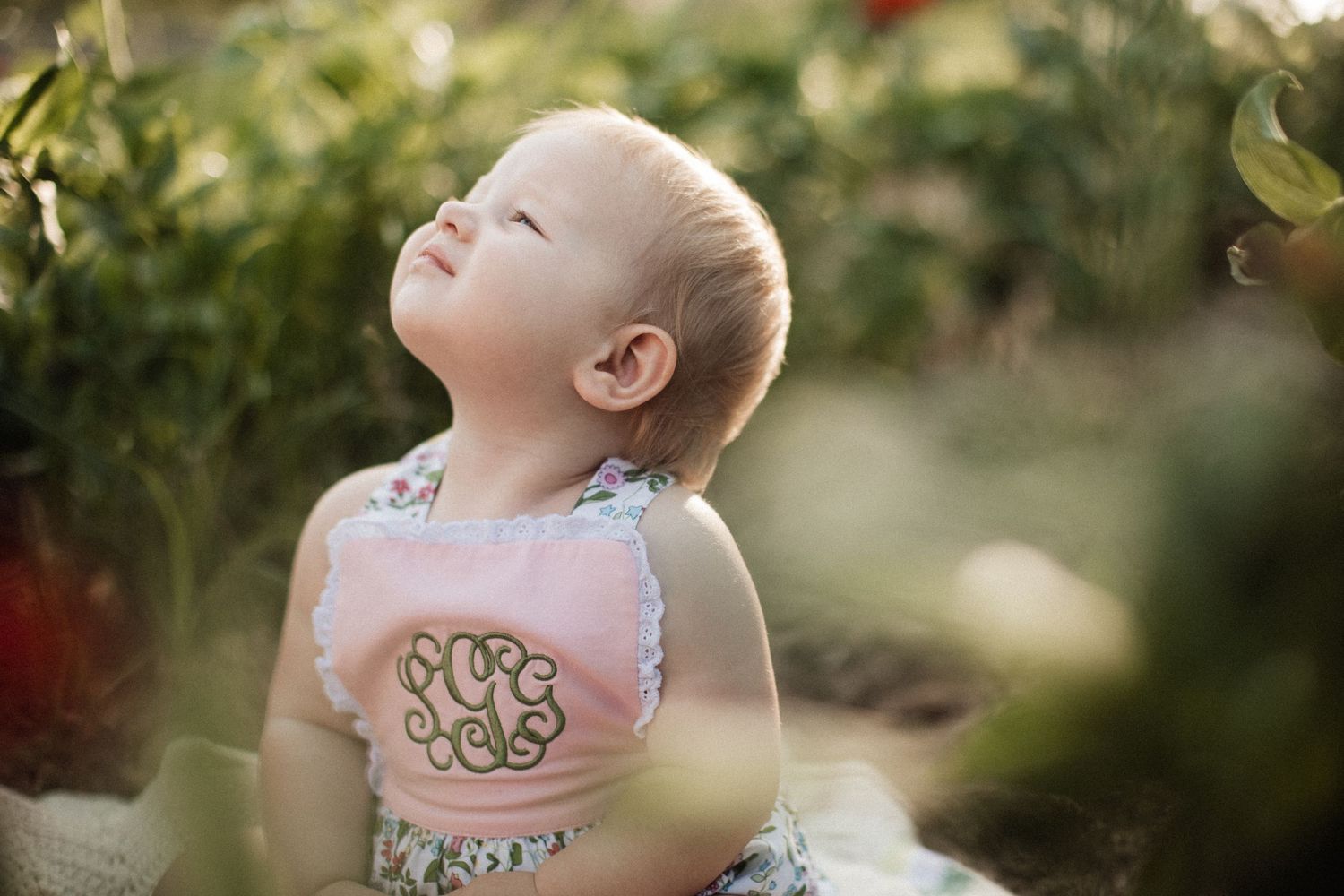 A baby in a pink dress explores a garden setting with blooming flowers and greenery on a sunny day.