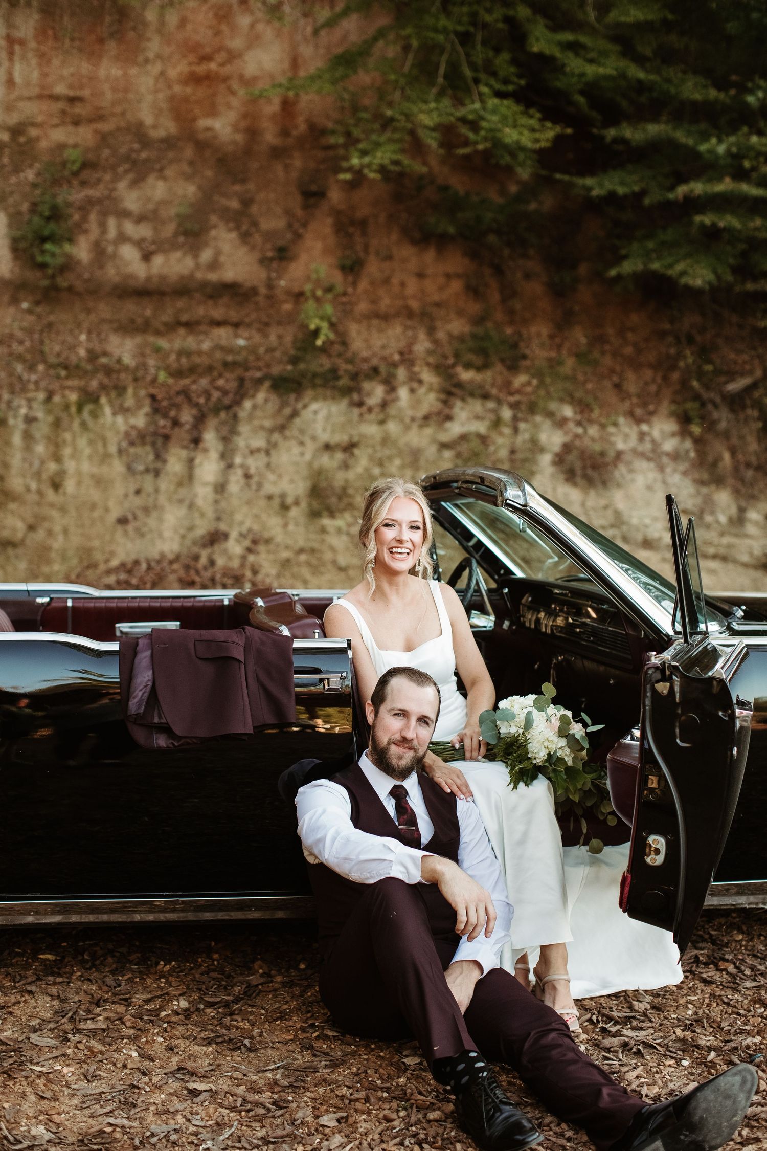 A couple shares a romantic moment in their wedding attire while sitting in a vintage car on a wooded path.