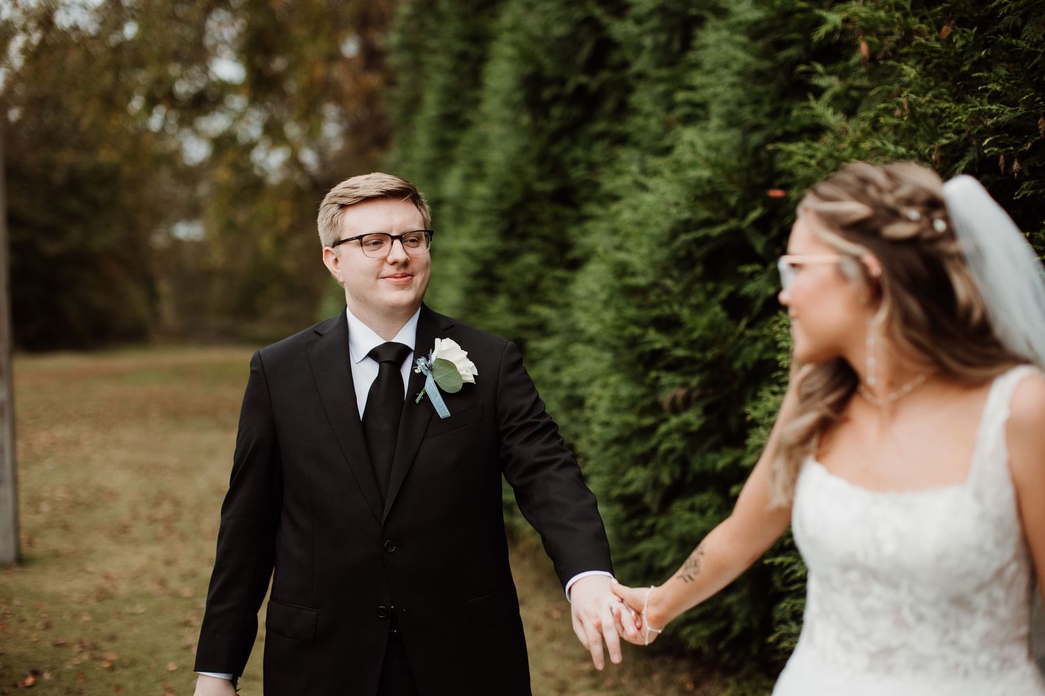 A couple in wedding attire hold hands while walking along a row of evergreen trees on their special day.