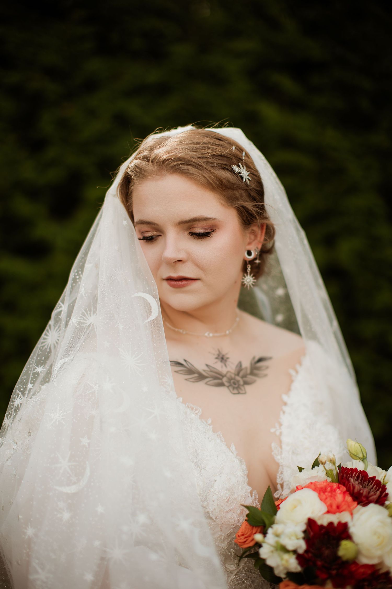 A bride in a white lace wedding dress with a veil holds a bouquet of red and white flowers.
