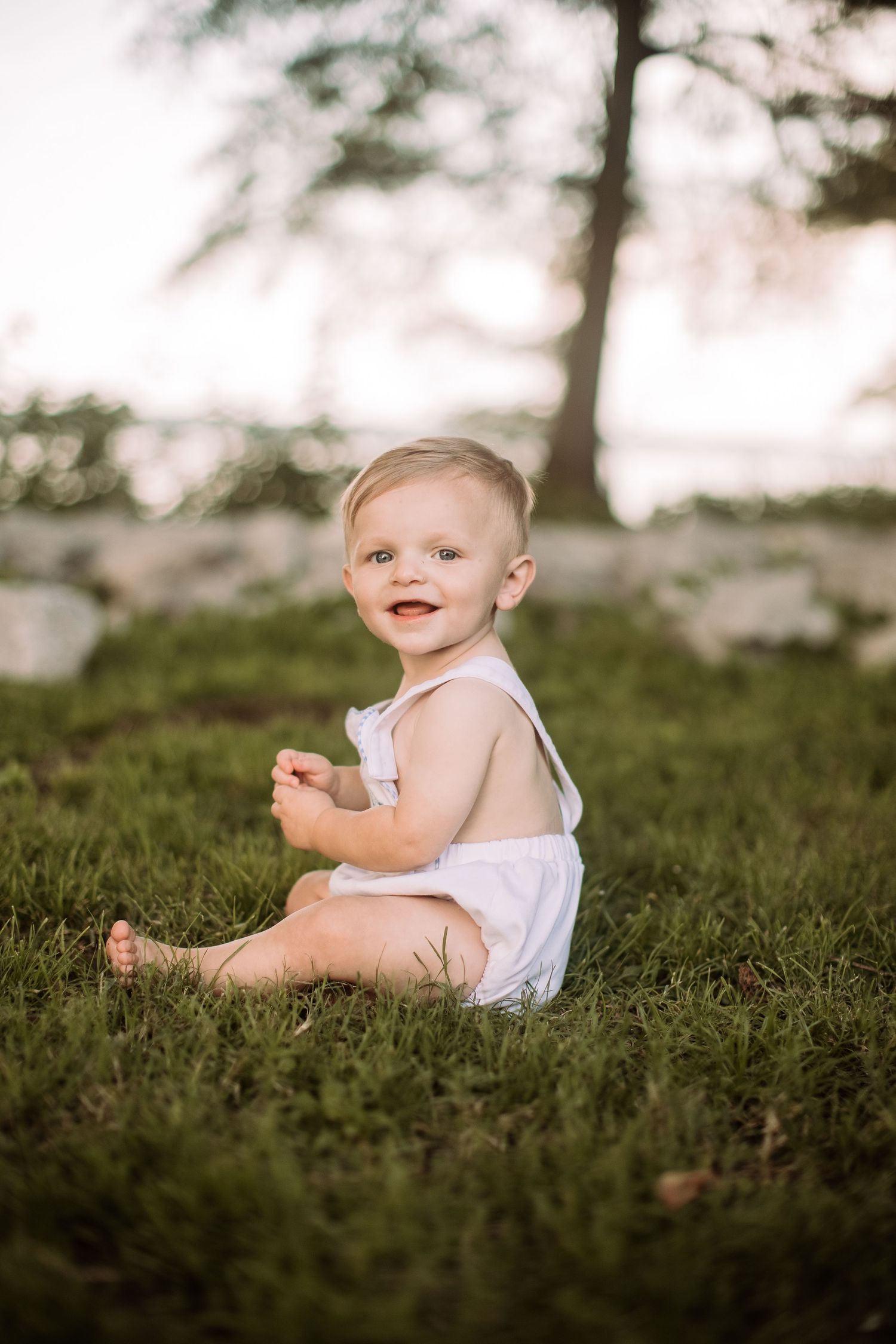 Small infant wearing white sits on green grass during golden hour lighting.
