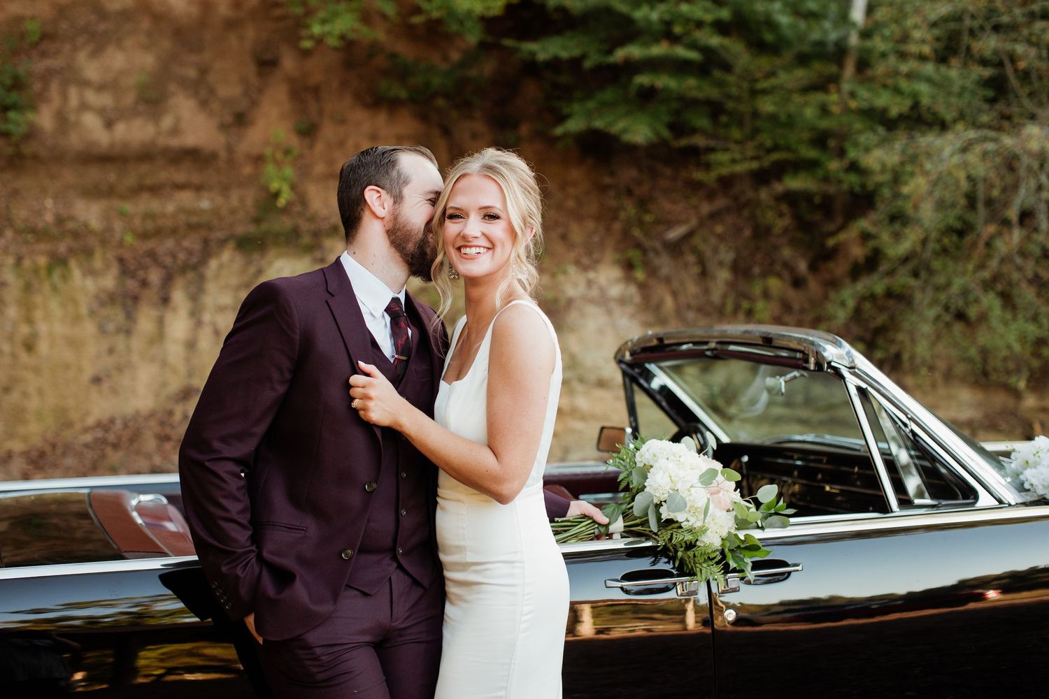 A romantic wedding couple poses with a vintage car in a lush forest setting during their special day.
