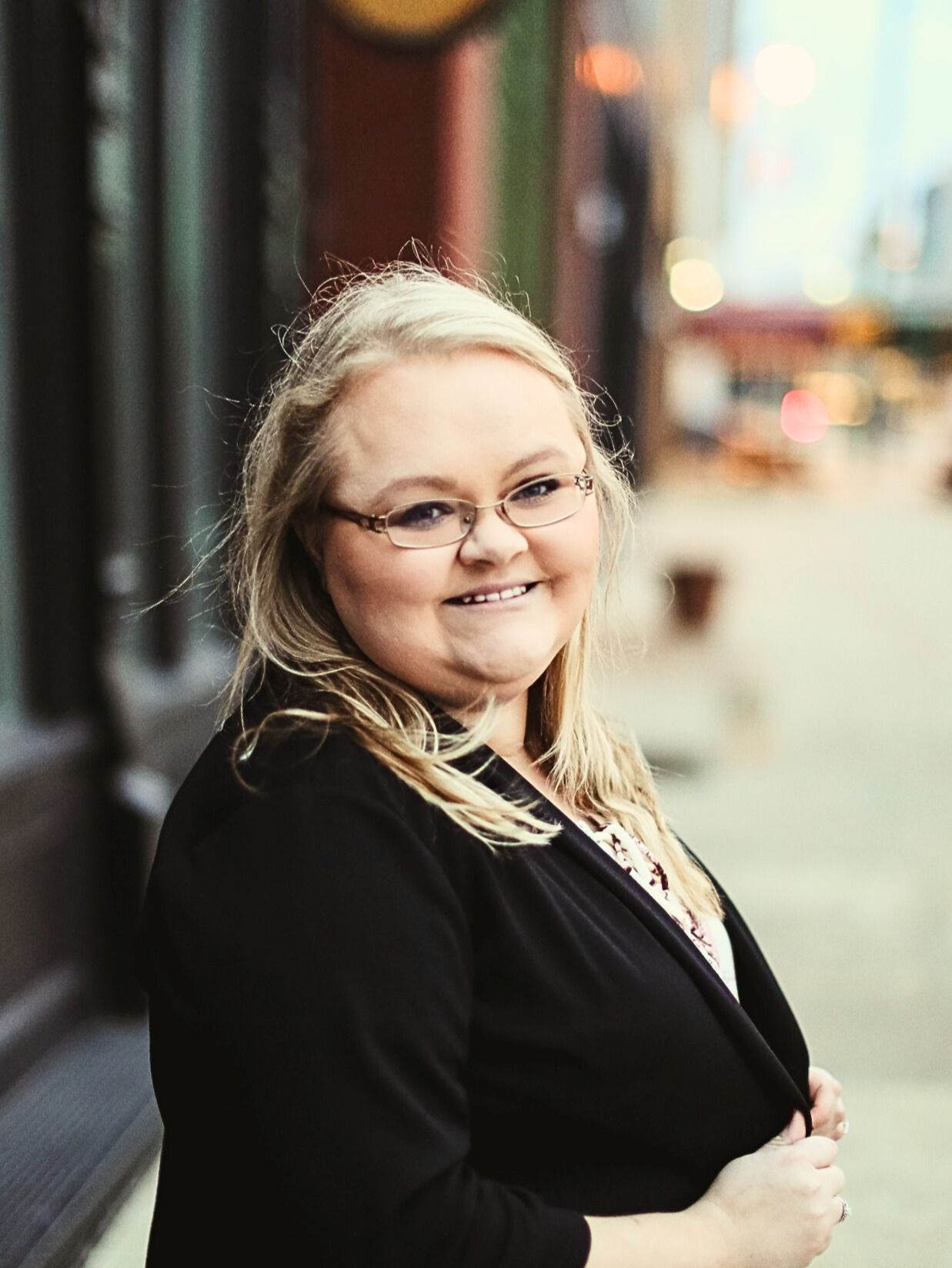 Professional headshot portrait of person in black blazer smiling warmly against blurred urban background.