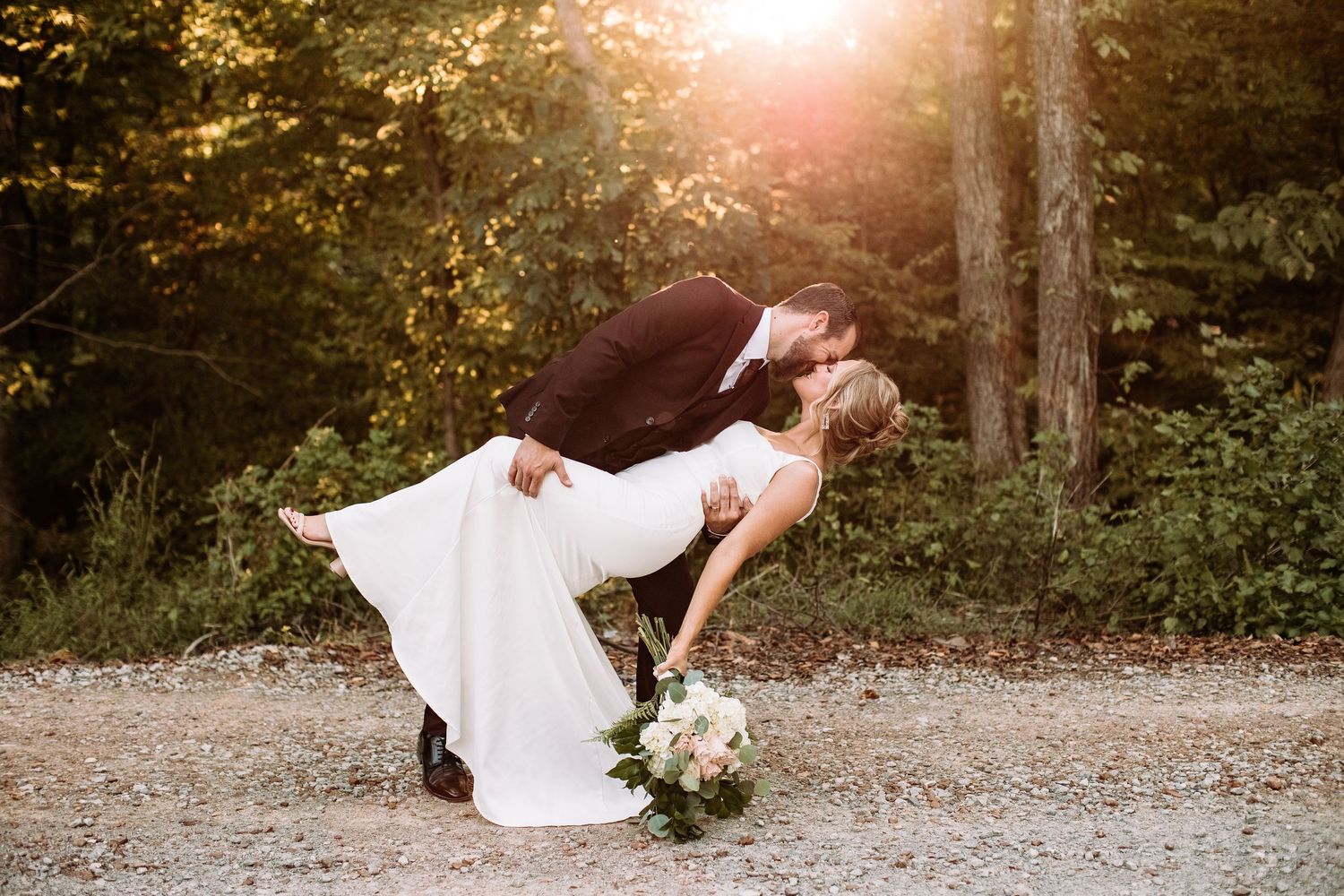A romantic dip pose on a sunlit forest path with a white wedding dress and bouquet of flowers.
