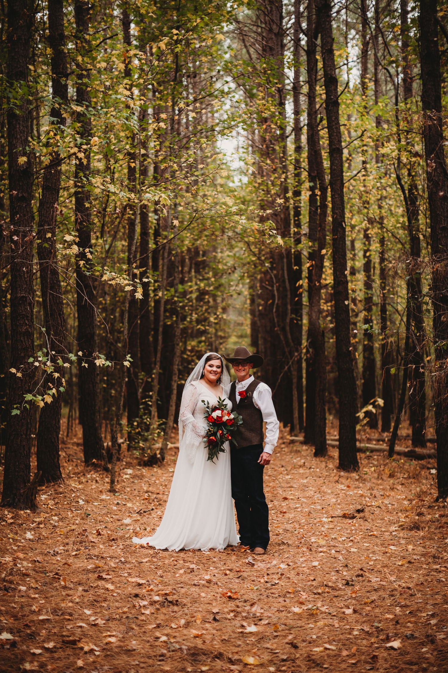 A couple in wedding attire stands on a path covered in fallen leaves surrounded by tall autumn trees in a forest.