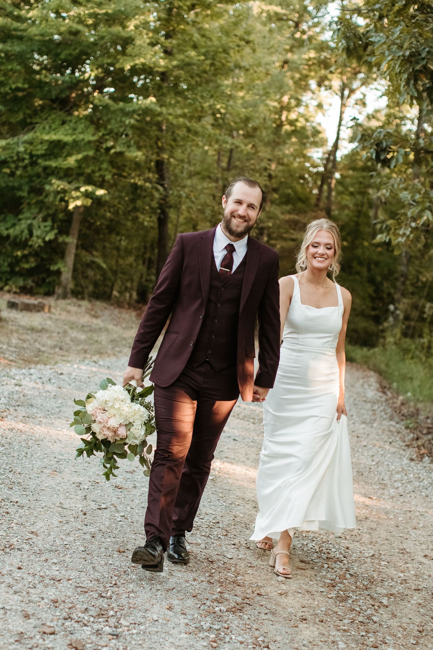 A couple in wedding attire walks together on a gravel path surrounded by trees during an outdoor autumn wedding.