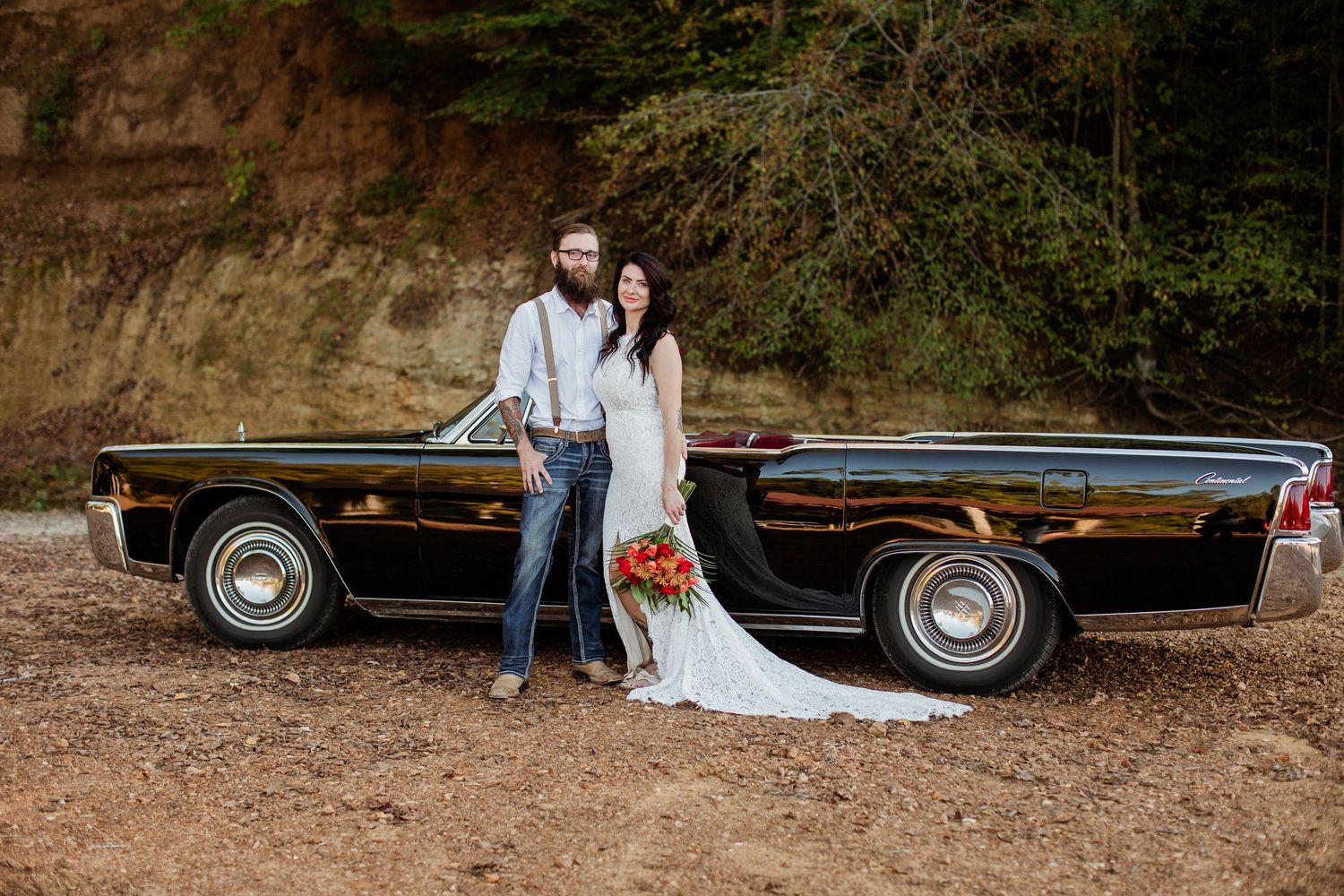 A classic black vintage car parked in a wooded area serves as a backdrop for a romantic photoshoot.