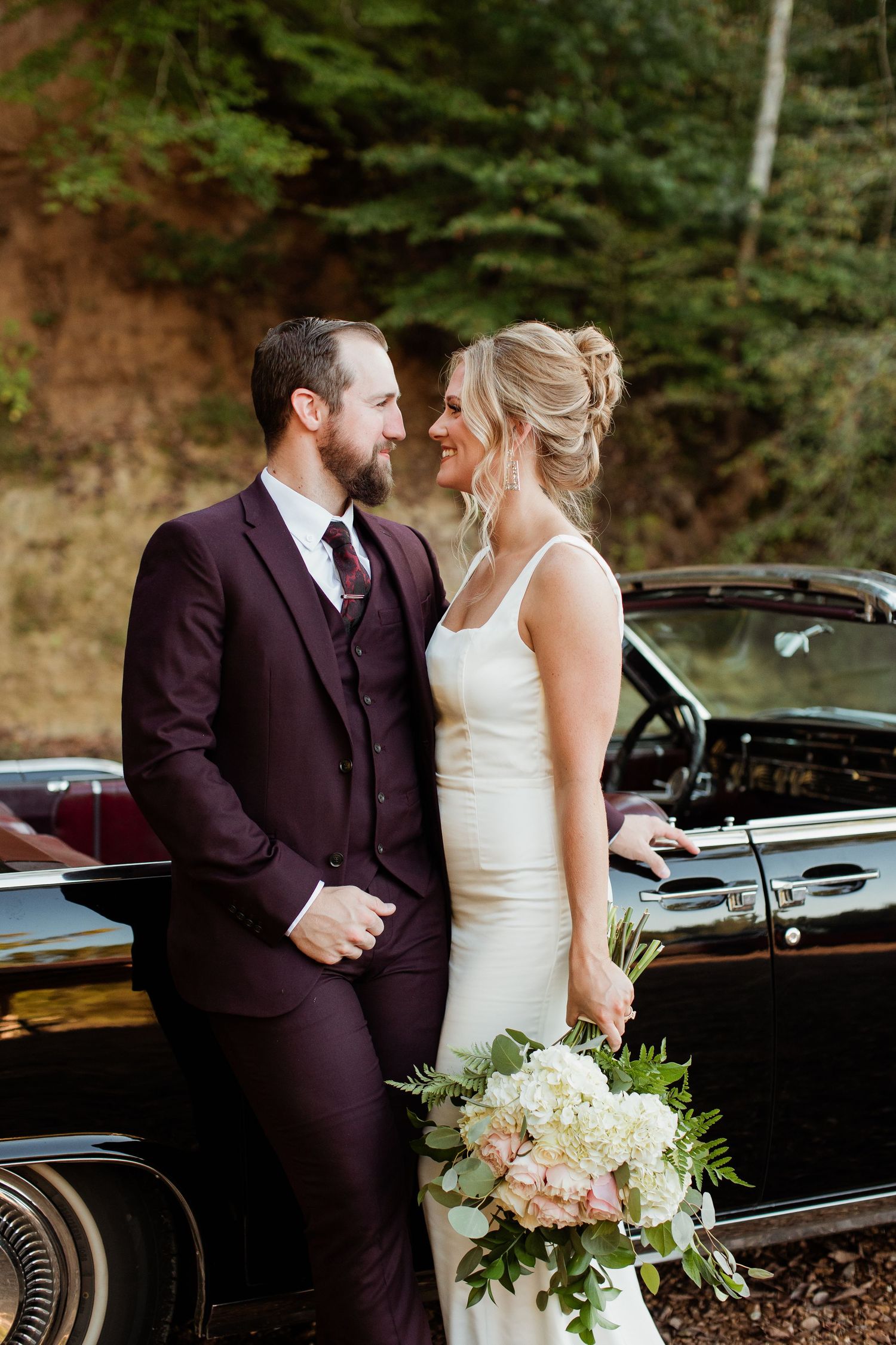 Newlywed couple shares an intimate moment beside a classic black car in a wooded outdoor setting.