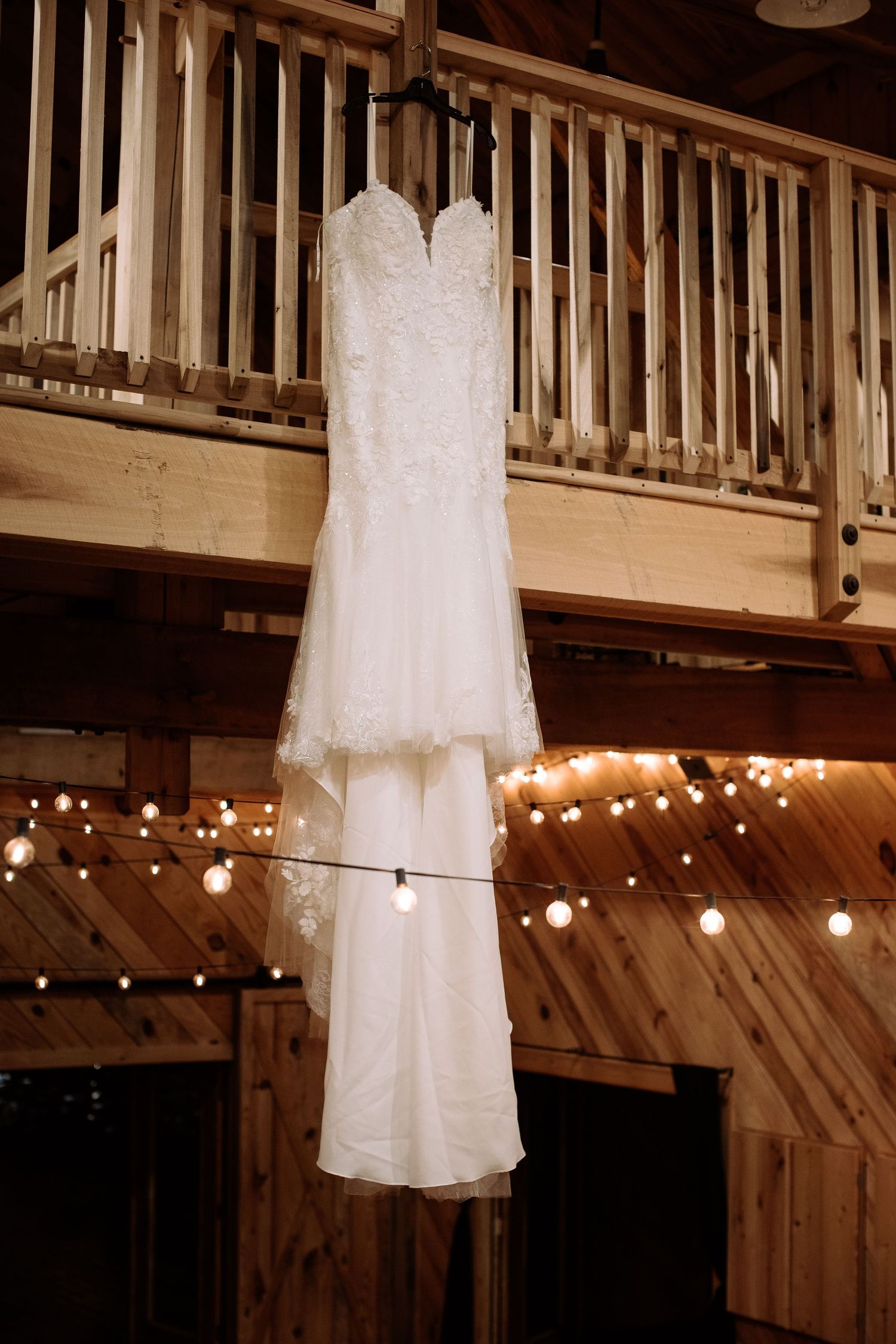 A white lace wedding dress hangs from wooden rafters in a rustic barn venue with string lights in the background.