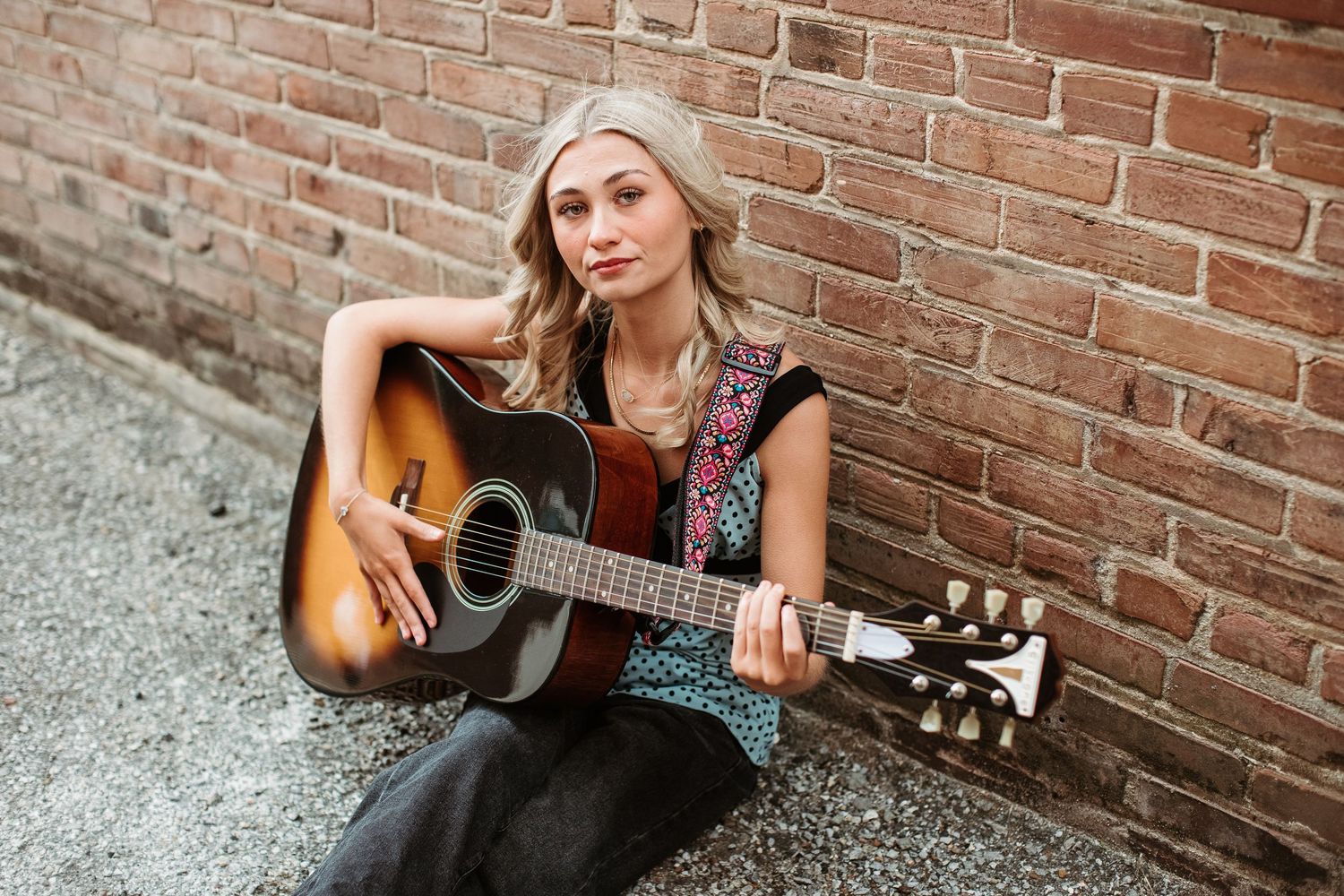 Young blonde woman sitting against brick wall playing acoustic guitar outdoors.