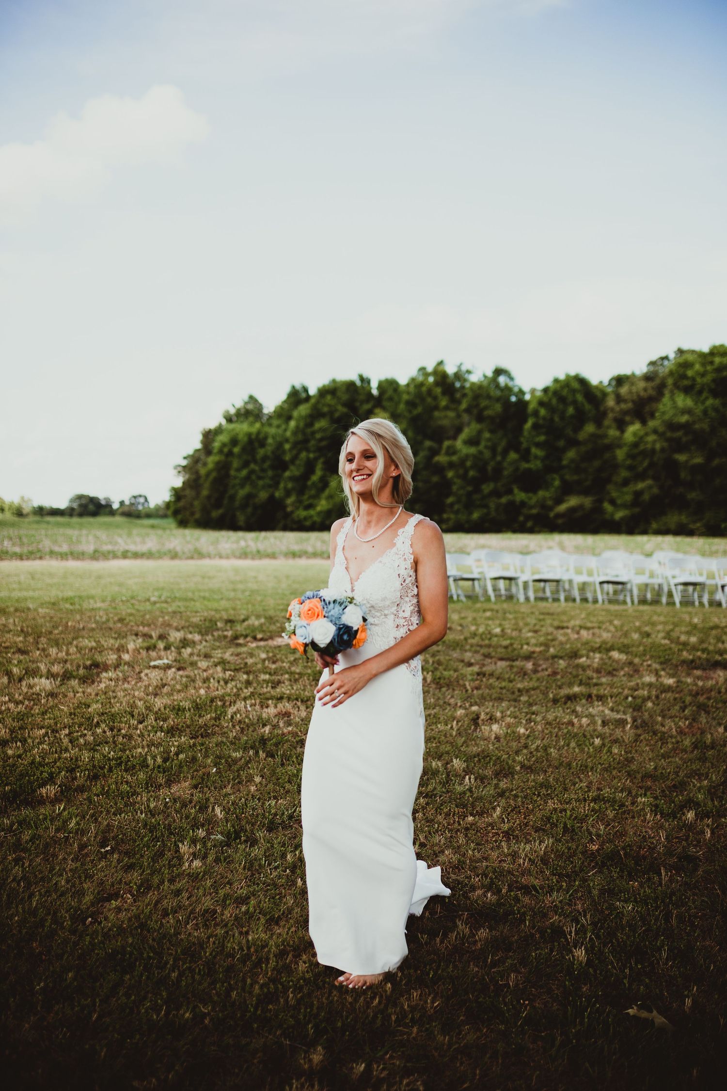 A bride in a white wedding dress walks through a rustic outdoor field with chairs set up for a ceremony in the distance.