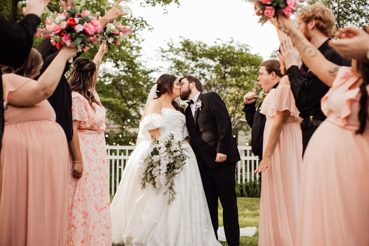 Wedding party in peach bridesmaid dresses celebrates as newlyweds share a romantic kiss in an outdoor garden setting.