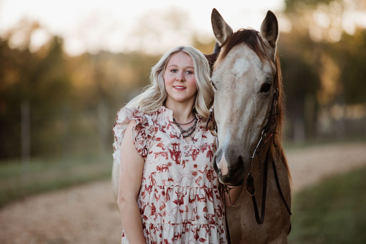 A woman in a floral dress stands with a brown horse in a golden-lit field at sunset.