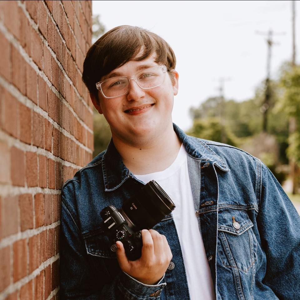 A photographer in a denim jacket smiles while holding a vintage camera against a brick wall background.
