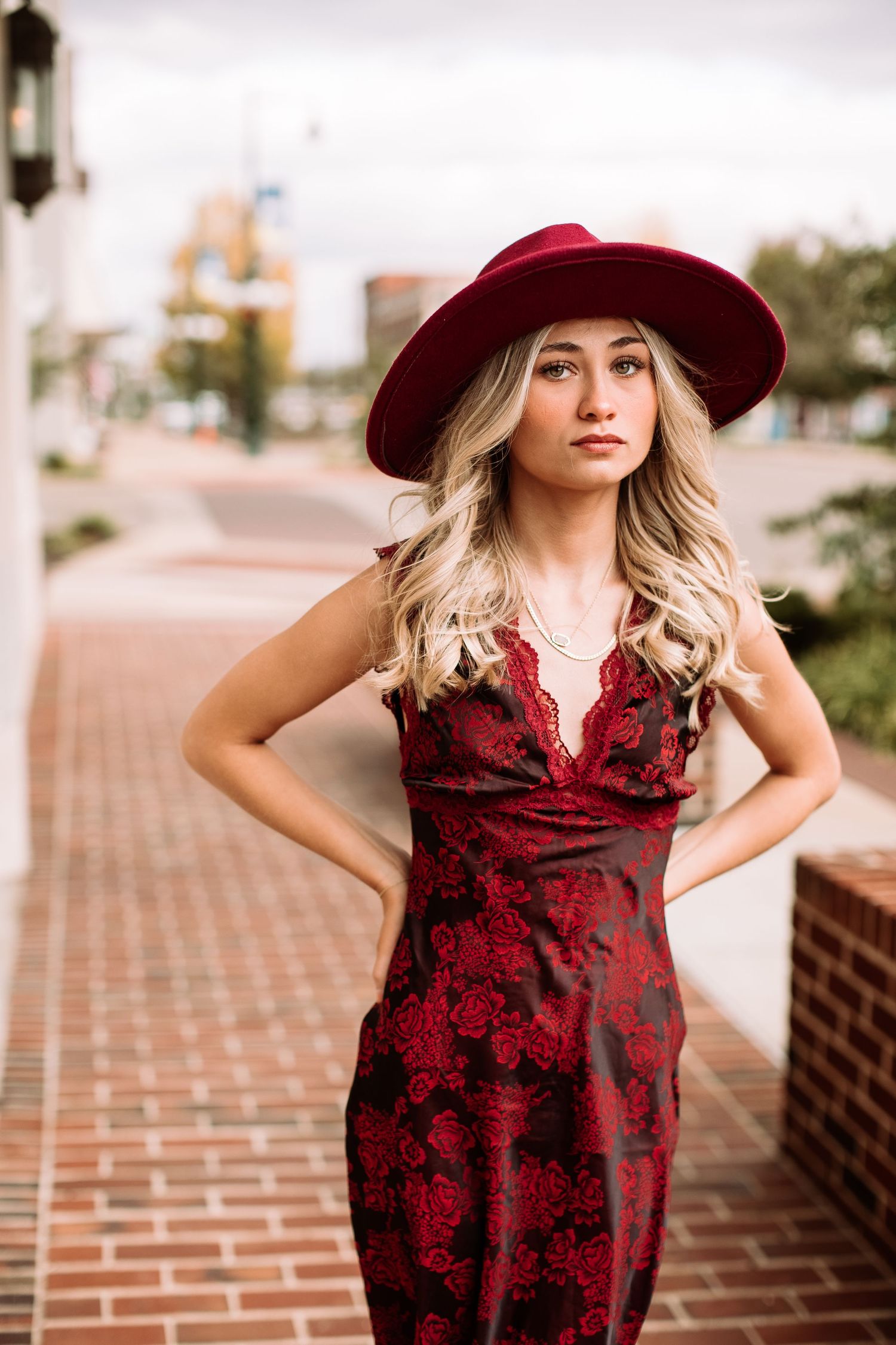 A burgundy floral dress paired with a wide-brimmed red hat creates an elegant autumn look against a brick backdrop.