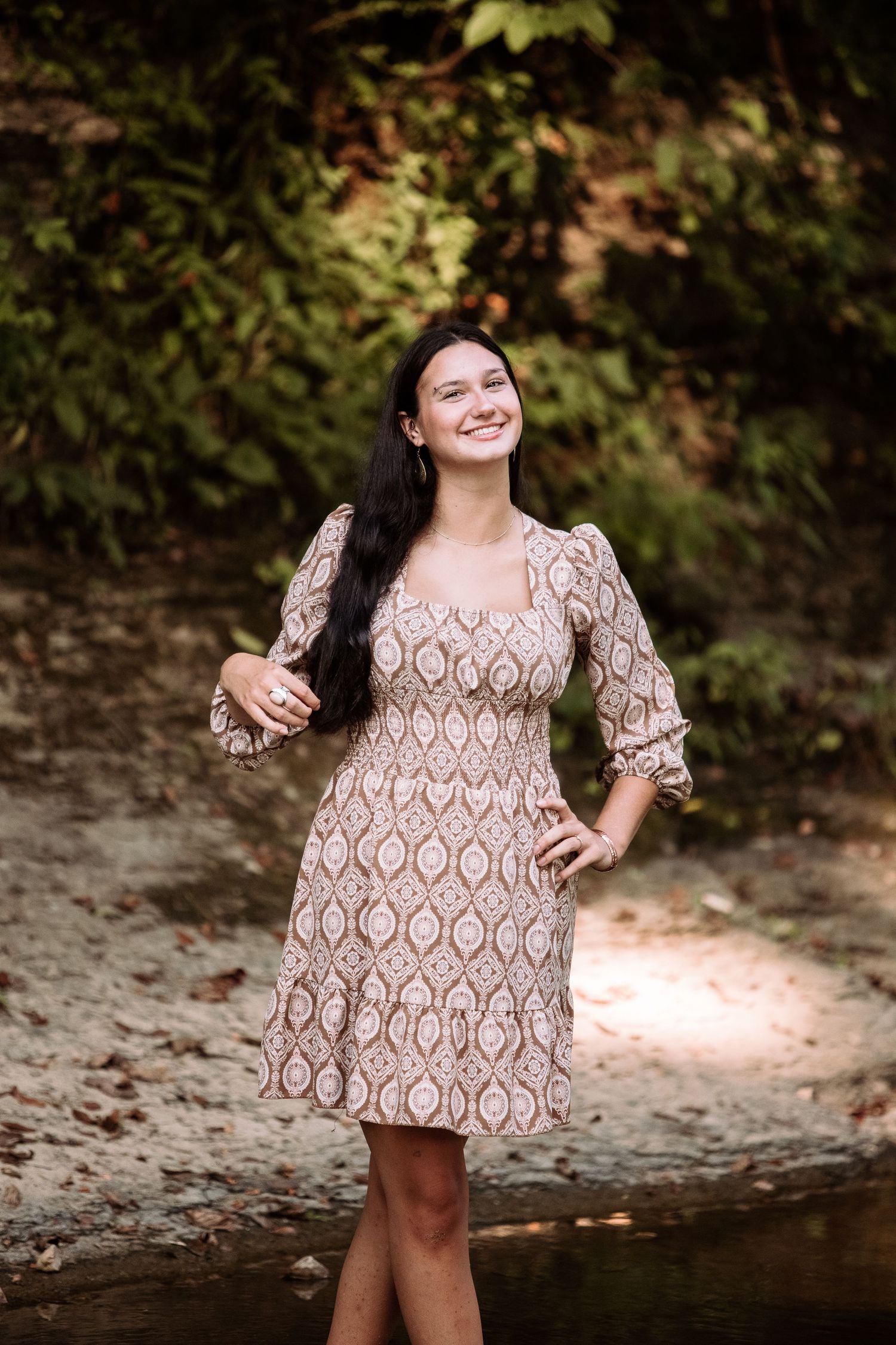 A person poses in a patterned babydoll dress by a creek in a natural outdoor setting.