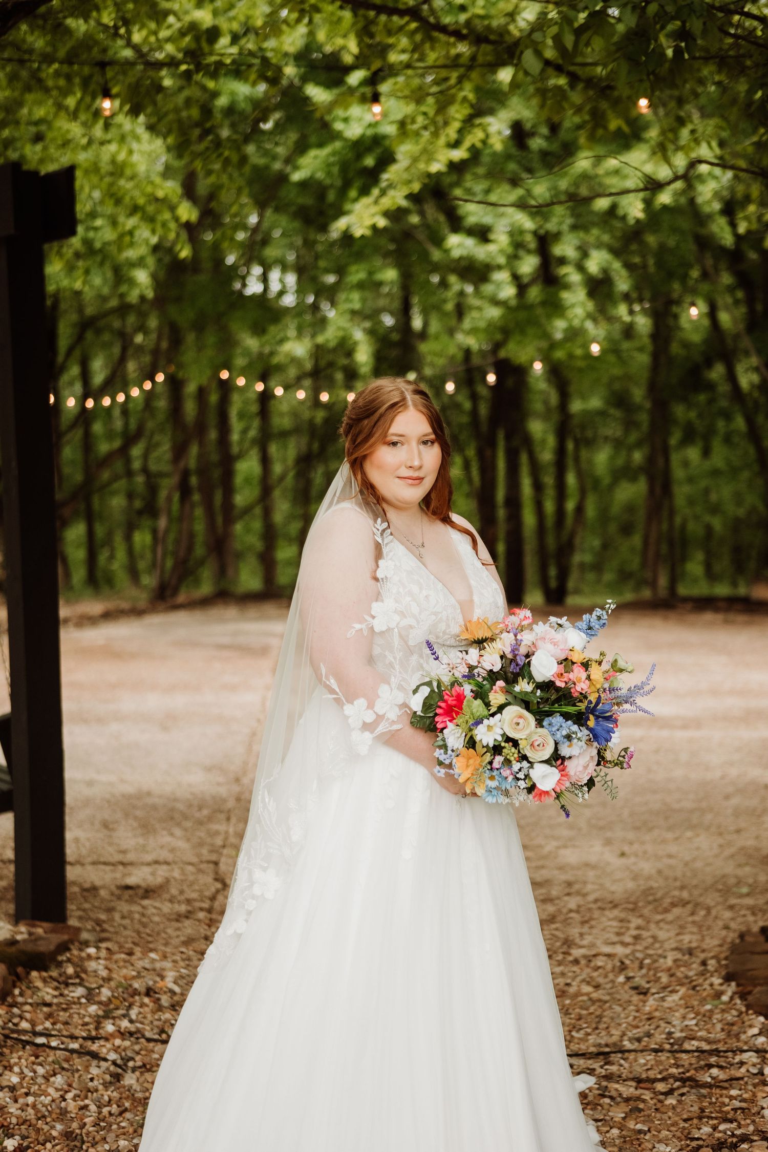 Bride in white lace gown holding colorful bouquet stands under string lights in a lush green forest setting.