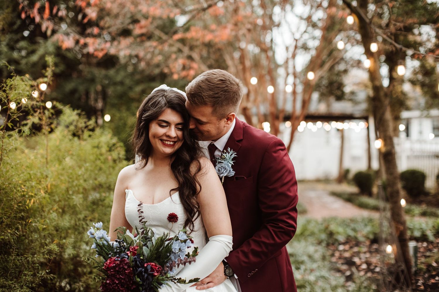 A couple in wedding attire embrace outdoors during autumn with fall foliage in the background.