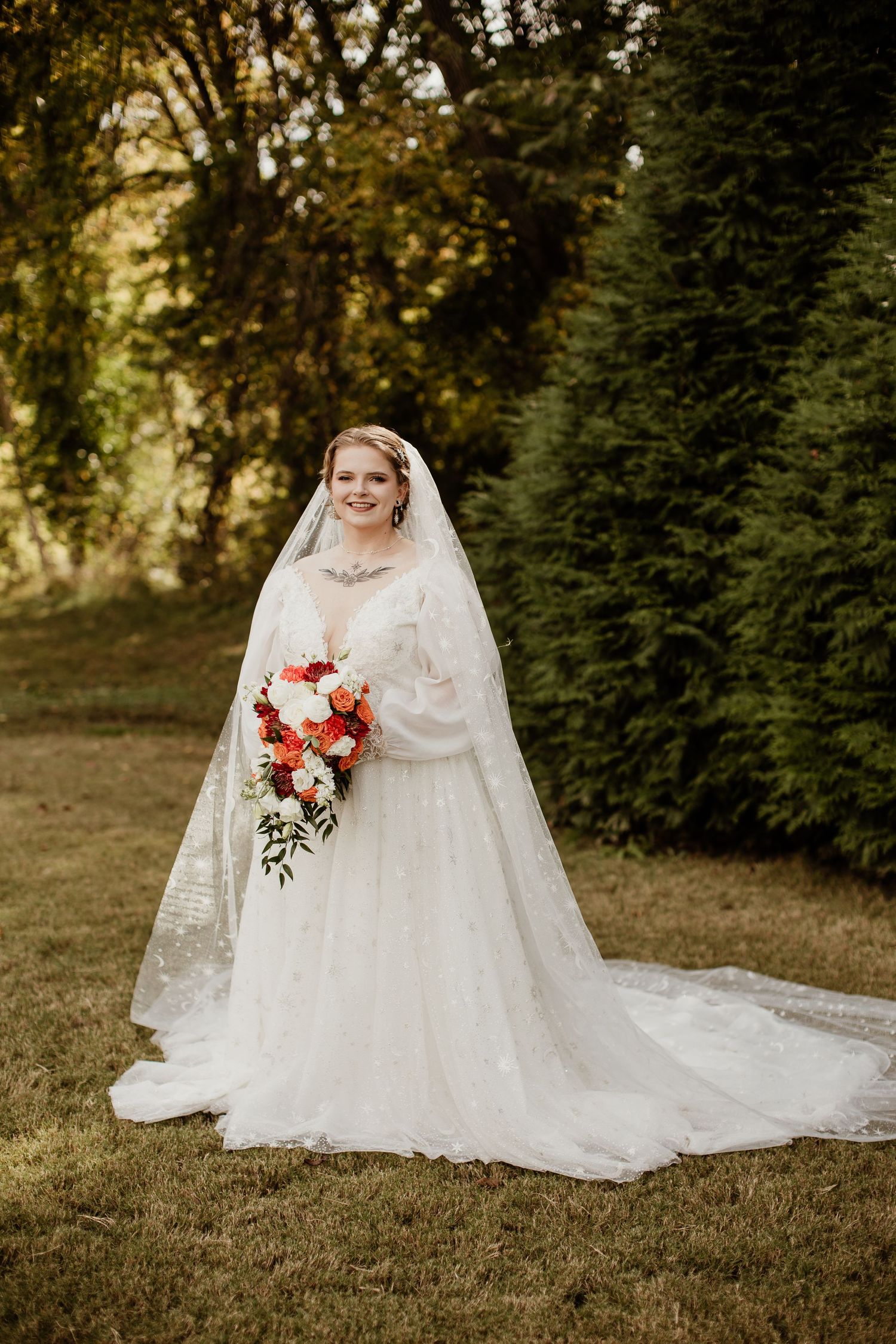 Bride in white lace ball gown and veil holding colorful bouquet, standing on green lawn with trees.