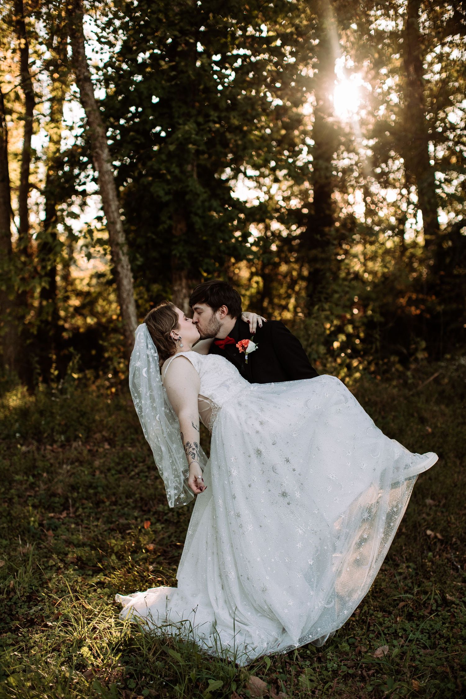 Groom carries bride in white lace gown while kissing in sunlit forest during golden hour wedding portrait.