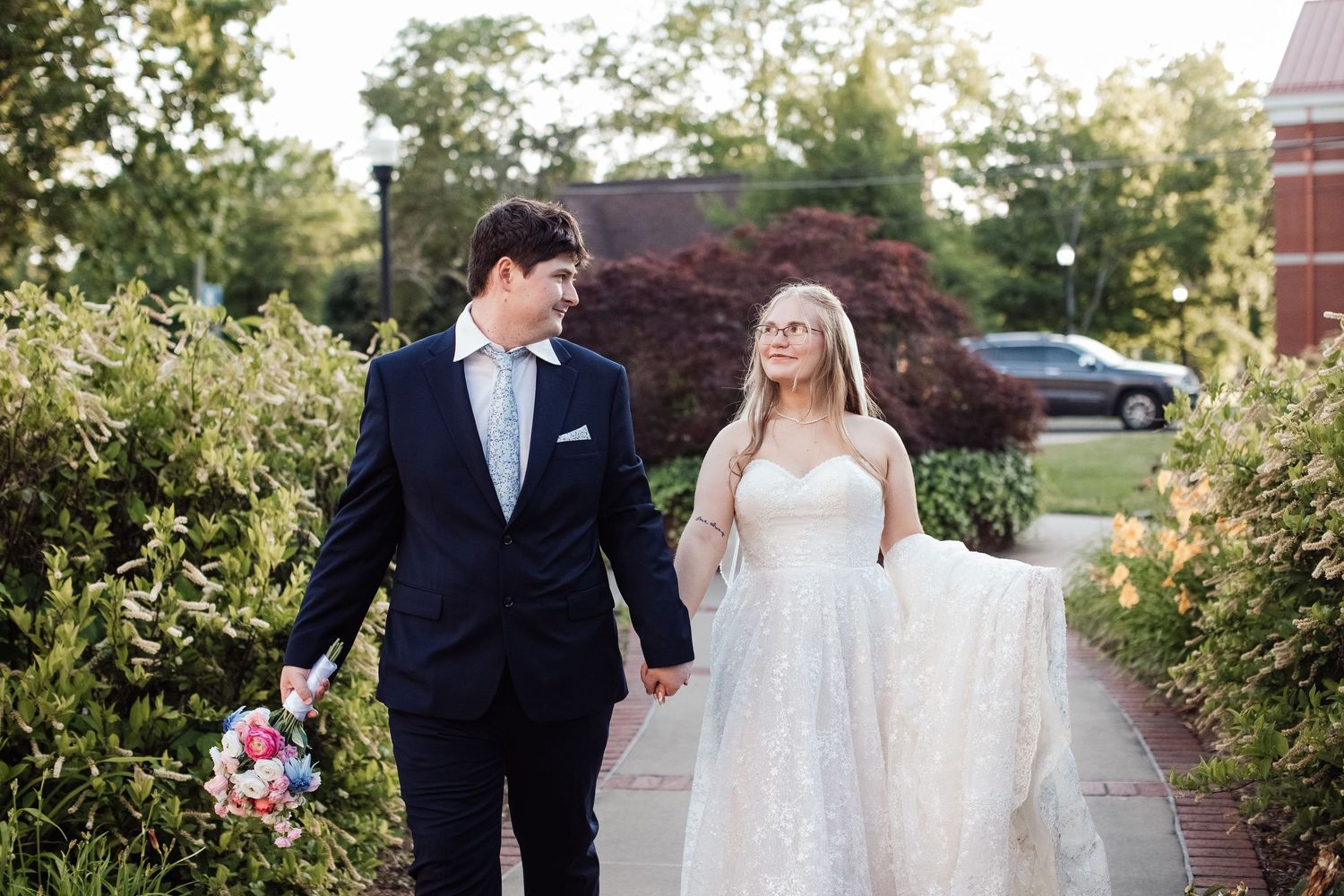 A bride and groom walk together along a tree-lined pathway during their outdoor wedding celebration.
