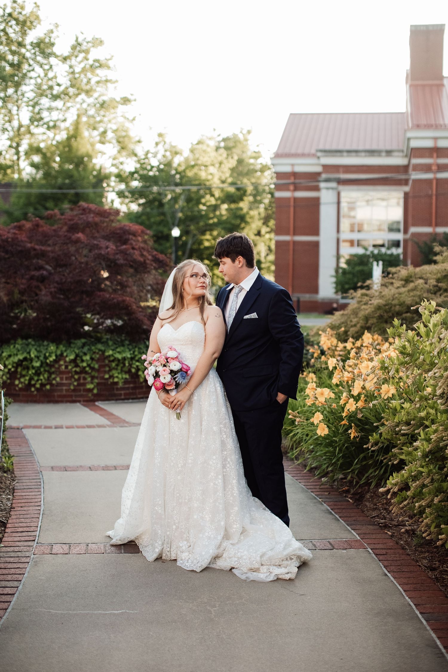 Romantic wedding couple shares tender moments on a garden pathway surrounded by blooming flowers and brick architecture.