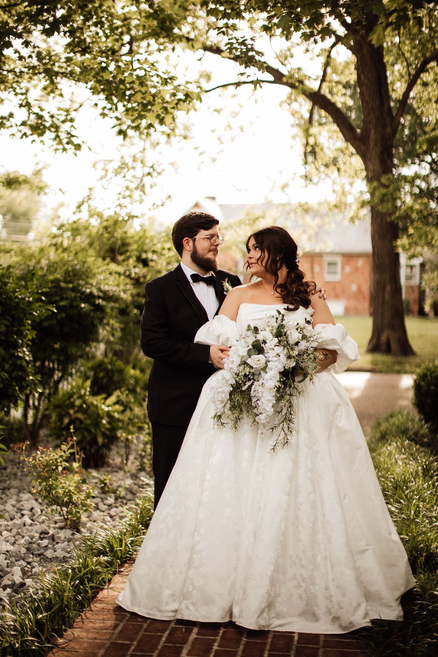 Newlywed couple in formal wedding attire stand together under a tree during golden hour on a brick pathway.
